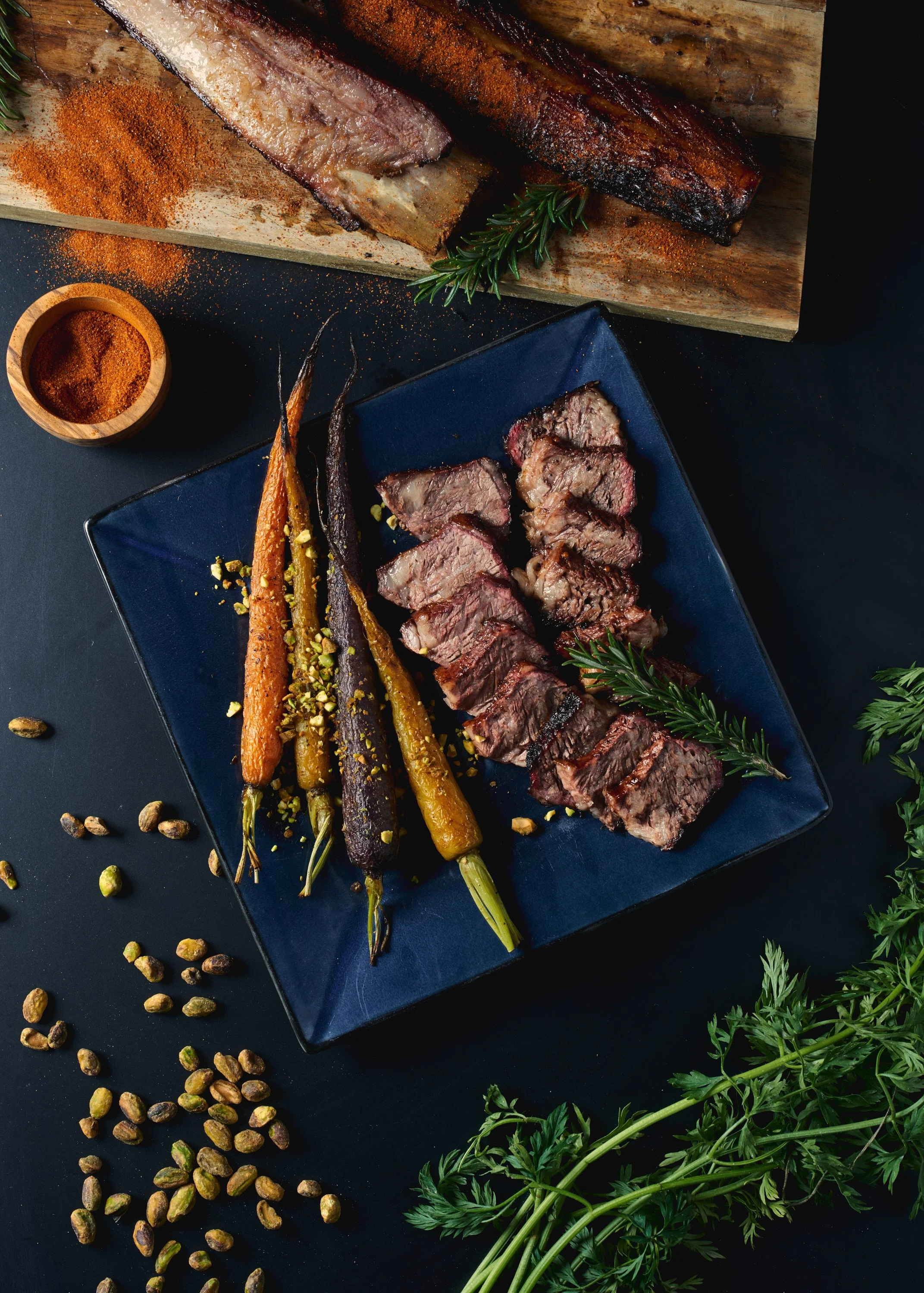 Sliced steak with roasted carrots and herbs on a dark plate, with a wooden serving board of grilled meat in the background, garnished with rosemary and sprinkled with crushed pistachios.