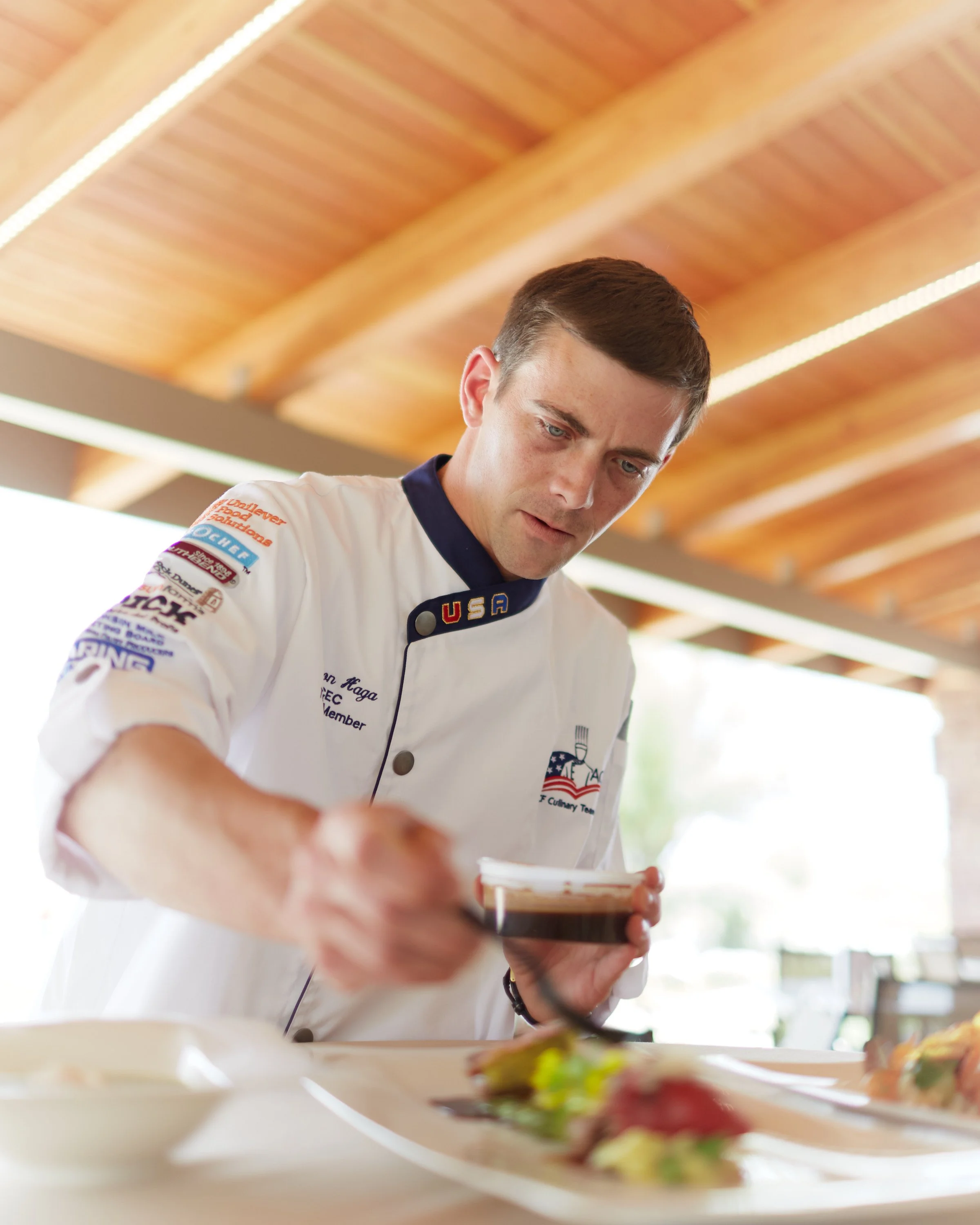 A male chef in a white uniform, with patches on the sleeve, is pouring sauce or dressing onto a plate of food. The background shows a wooden ceiling.