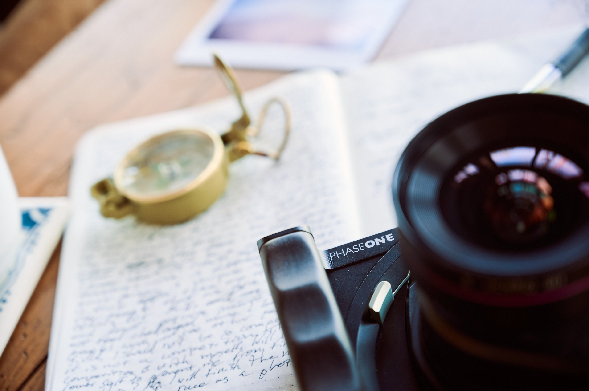 Close-up of a camera, an open notebook with handwritten notes, a pair of glasses, and a smartphone on a wooden surface.