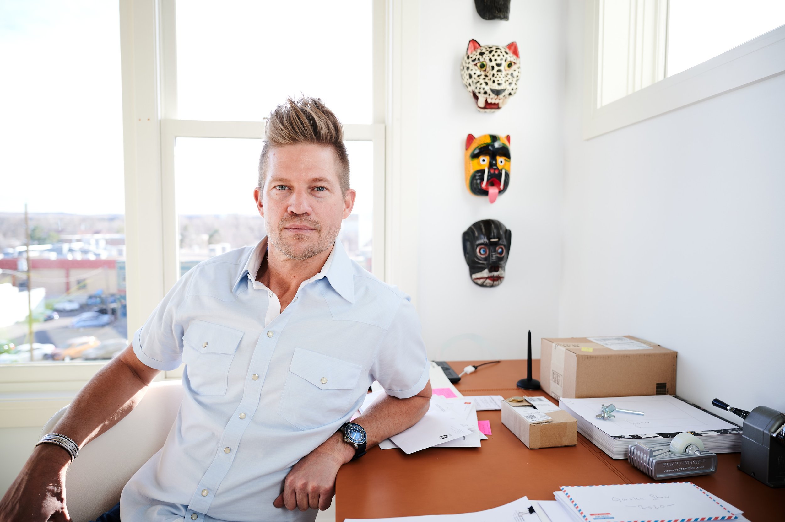 Man with blonde hair and beard sitting at a cluttered desk in a bright office, with colorful decorative masks on the wall behind him.