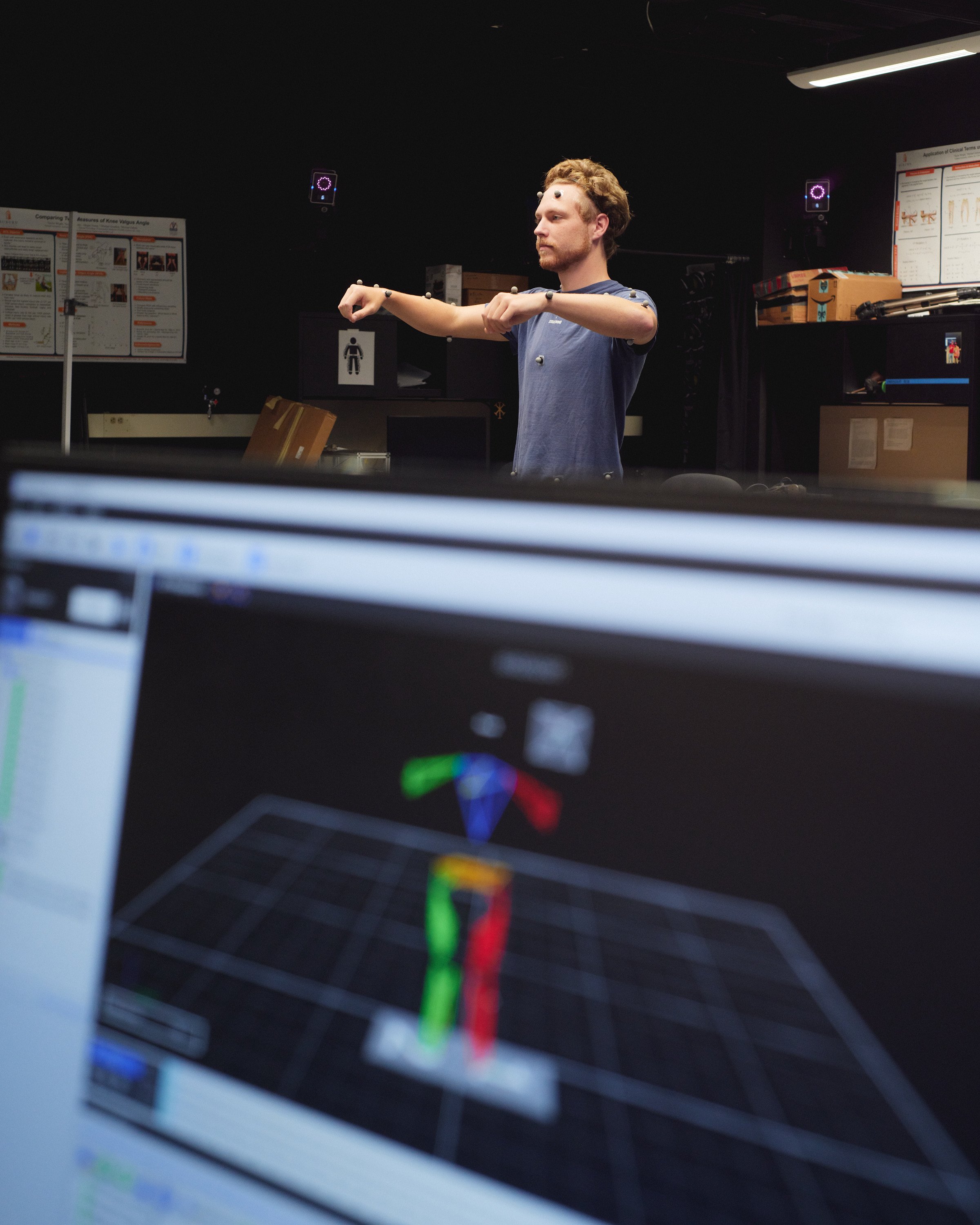 A man standing in a dark room with sensors attached to his arms and face, conducting motion capture testing, with a computer screen displaying a 3D model of his movement in the foreground.