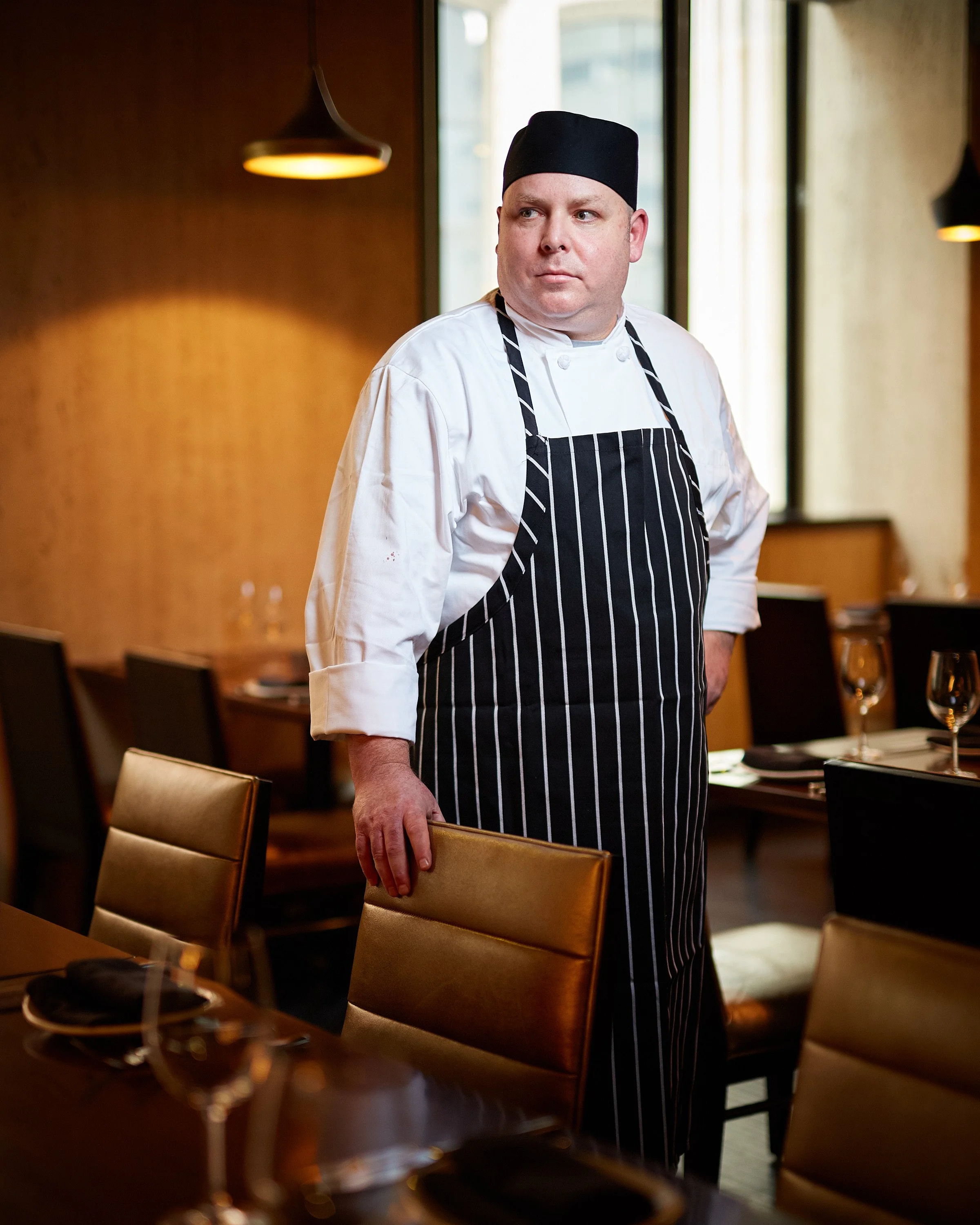 A male chef in a black and white striped apron and black hat standing in a restaurant dining area, resting his hand on a chair.