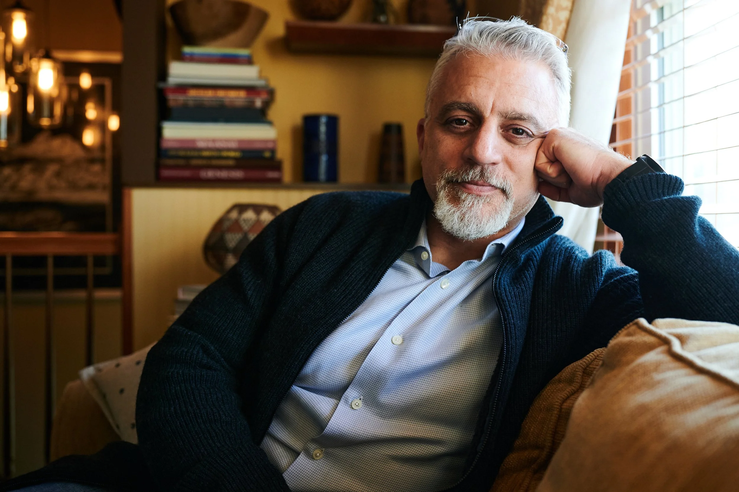 An older man with white hair and a beard, sitting on a couch, resting his head on his hand near a window with blinds, in a warmly lit room with bookshelves and decor.