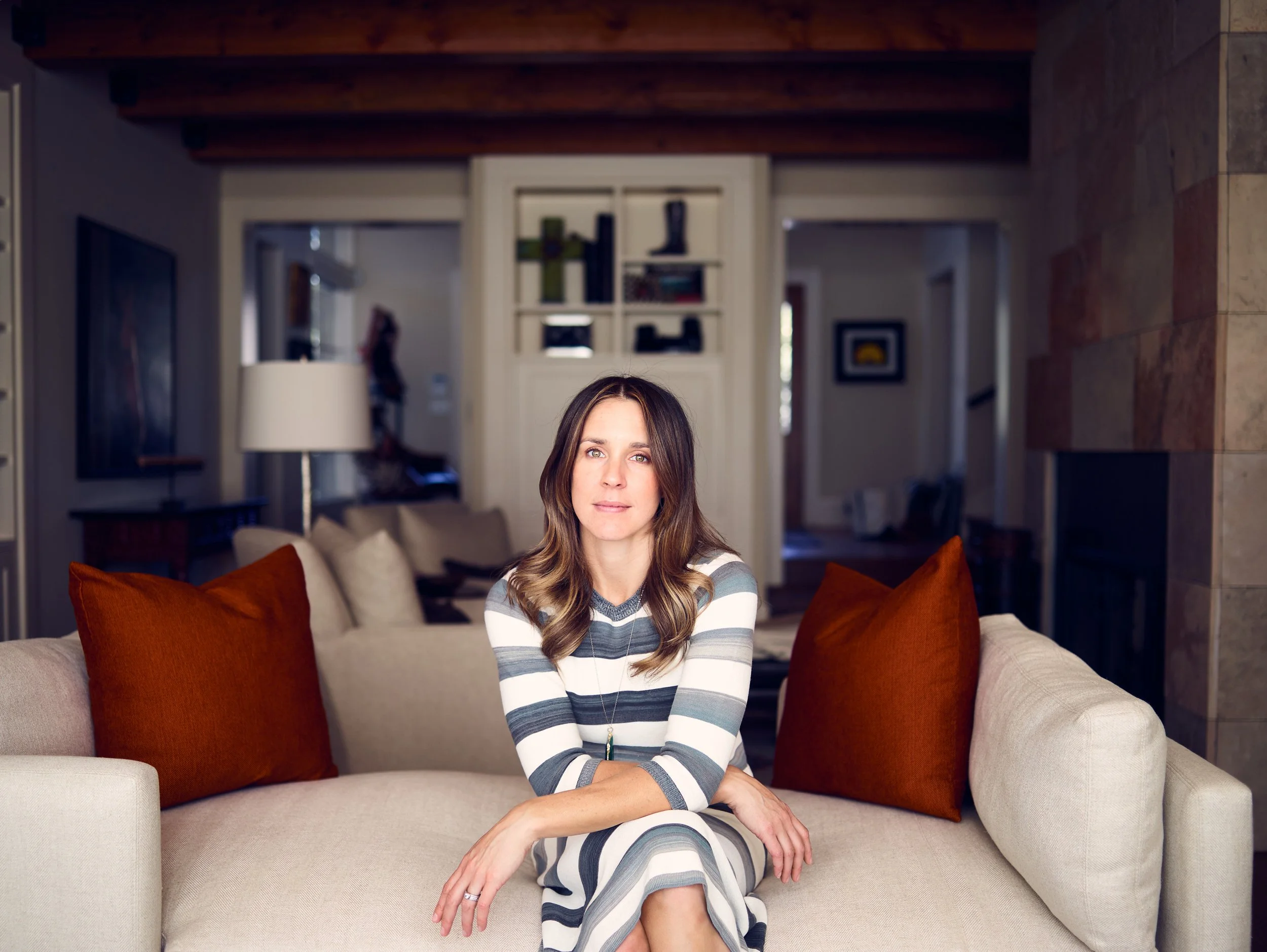 A woman sitting on a beige sofa with orange pillows in a living room with wooden ceiling beams and a stone wall on the right.