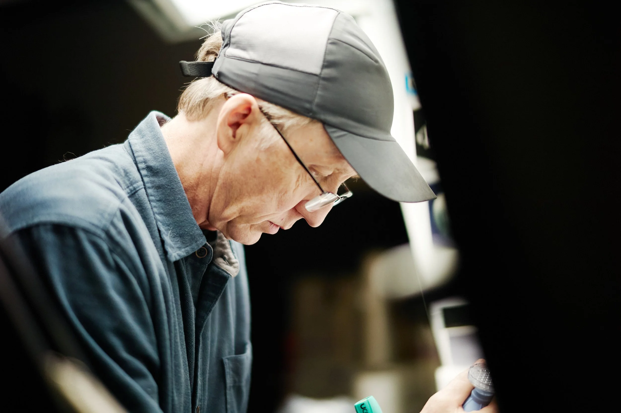 A man wearing a gray cap, glasses, and a blue jacket is looking down and reading something in his hand.