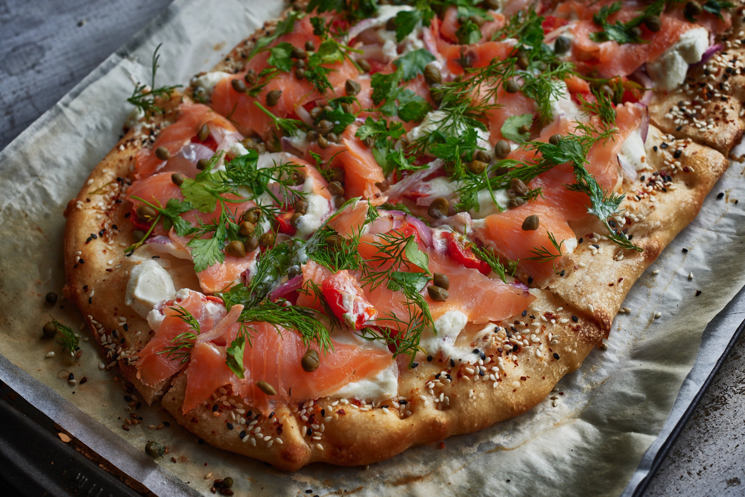 A pizza topped with smoked salmon, fresh herbs, capers, and cheese on a baking sheet lined with parchment paper.
