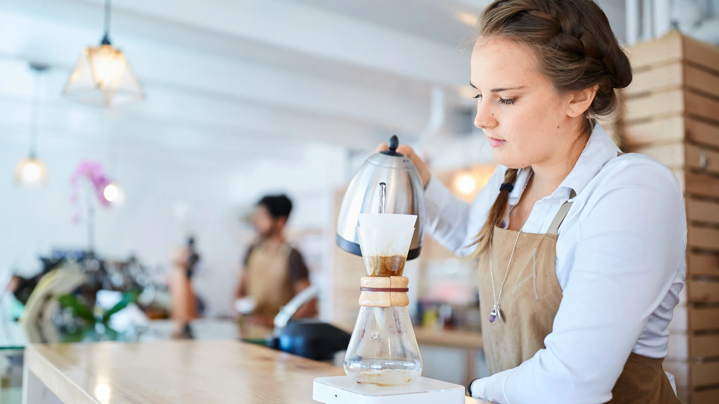 Young woman making pour-over coffee in a café, wearing a white shirt and beige apron, with a blurred background of other café staff and customers.