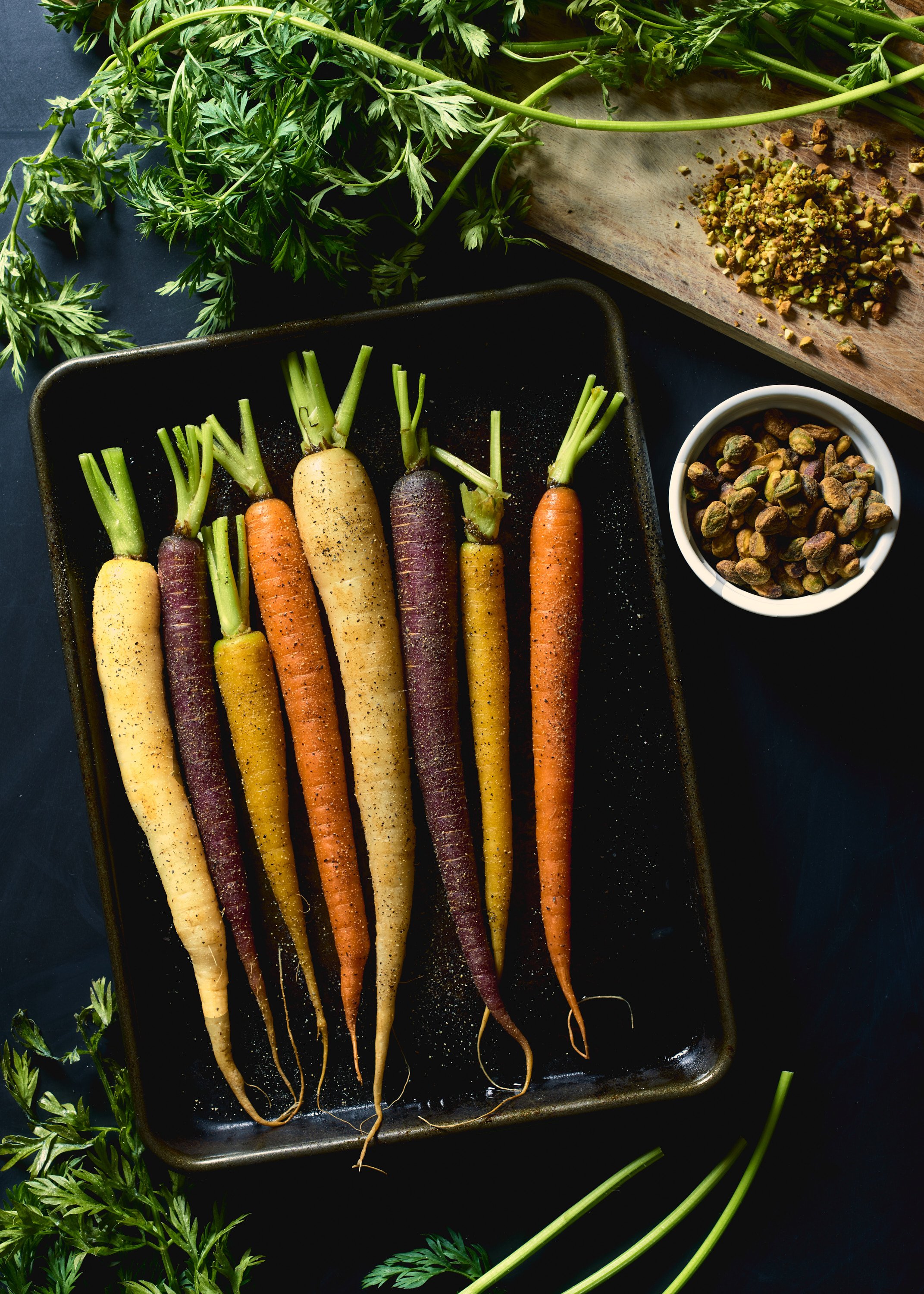 Colorful heirloom carrots with green tops on a black baking tray, surrounded by herbs, chopped pistachios, and cardamom pods in bowls.