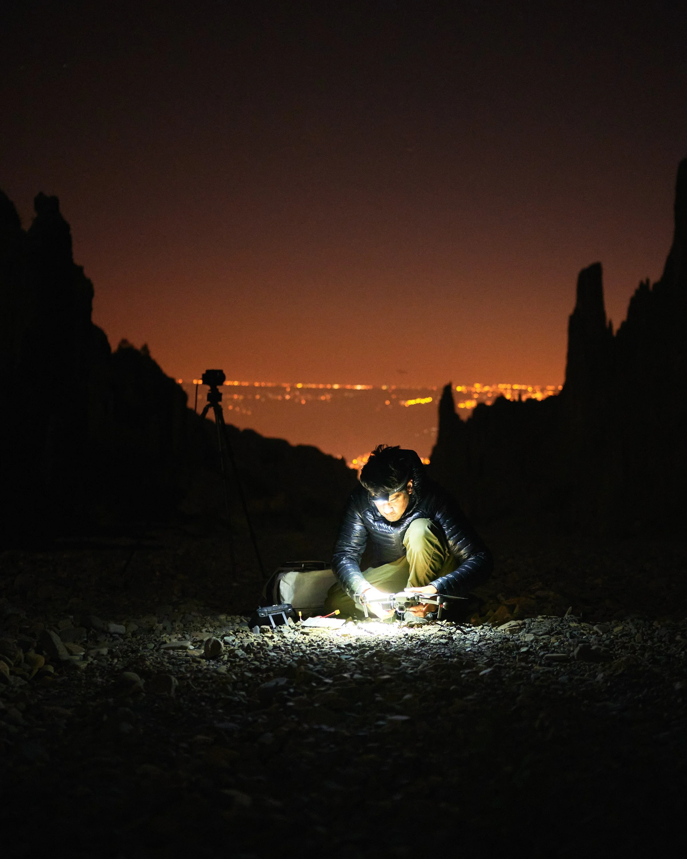 Person crouching on rocky ground at night, working with equipment illuminated by a flashlight. Skyline with city lights visible in the distance, rocky formations framing the scene.