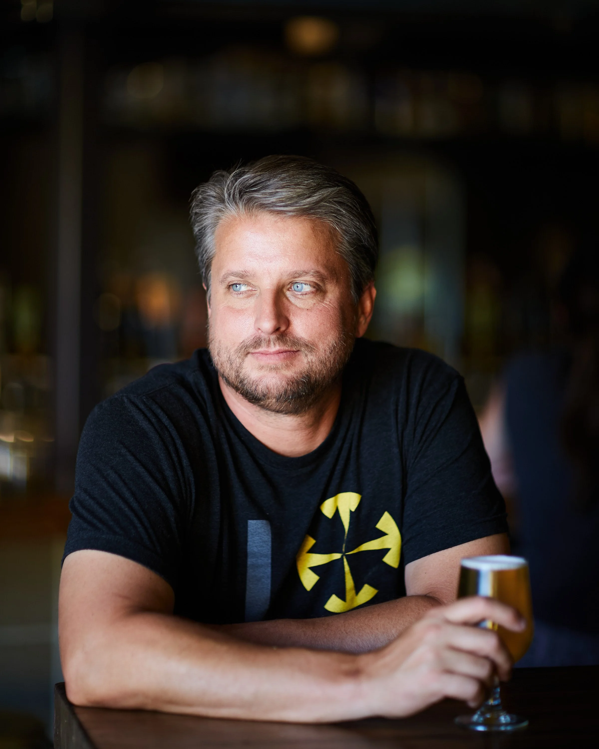 A middle-aged man with gray hair and a beard, wearing a black t-shirt with a yellow design, sitting at a table in a dimly lit space, holding a glass of beer, looking to the side.