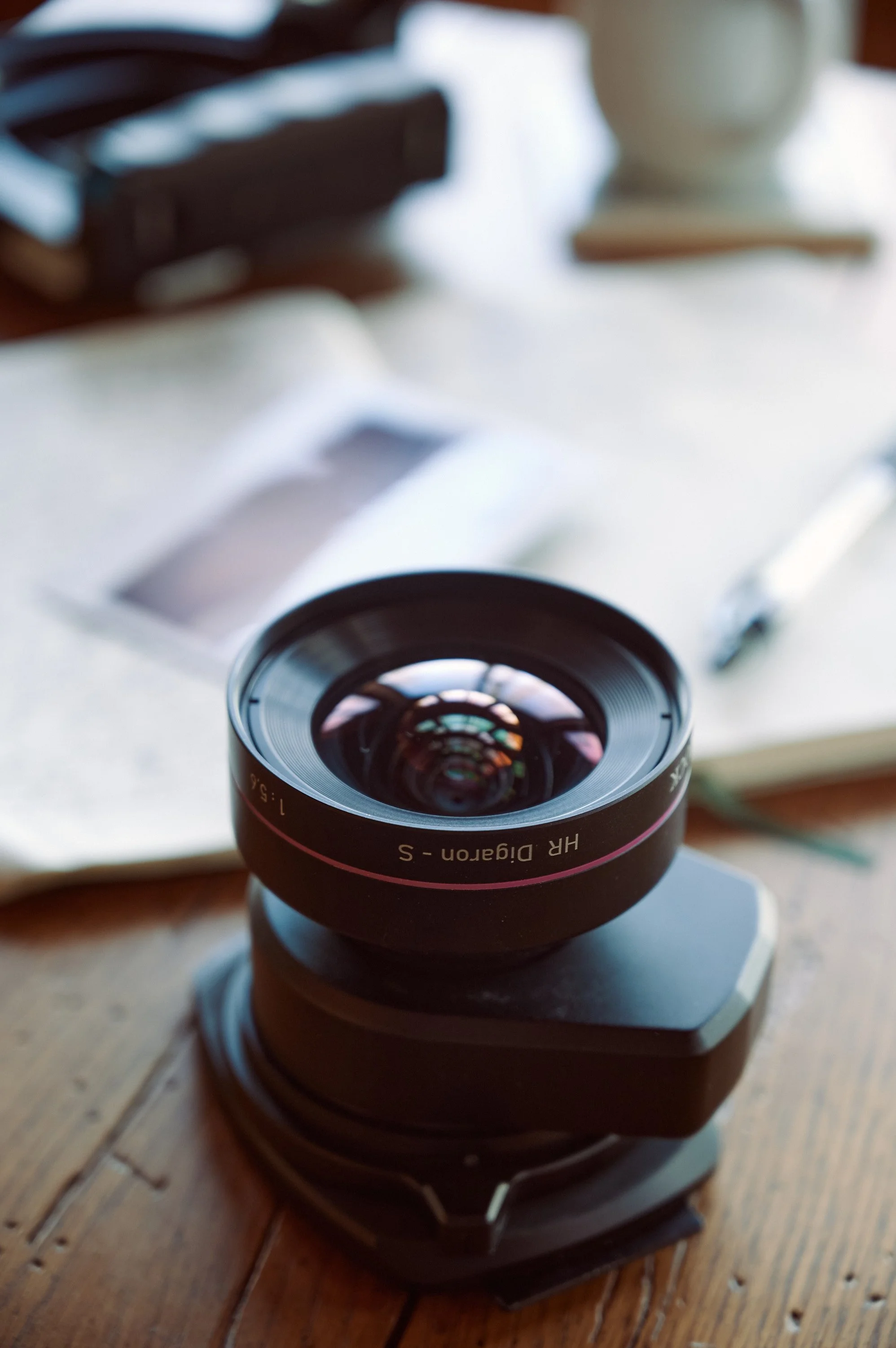Close-up of a camera lens labeled 'HR Diagon S' on a wooden table with blurred background of papers, pen, and a lamp.