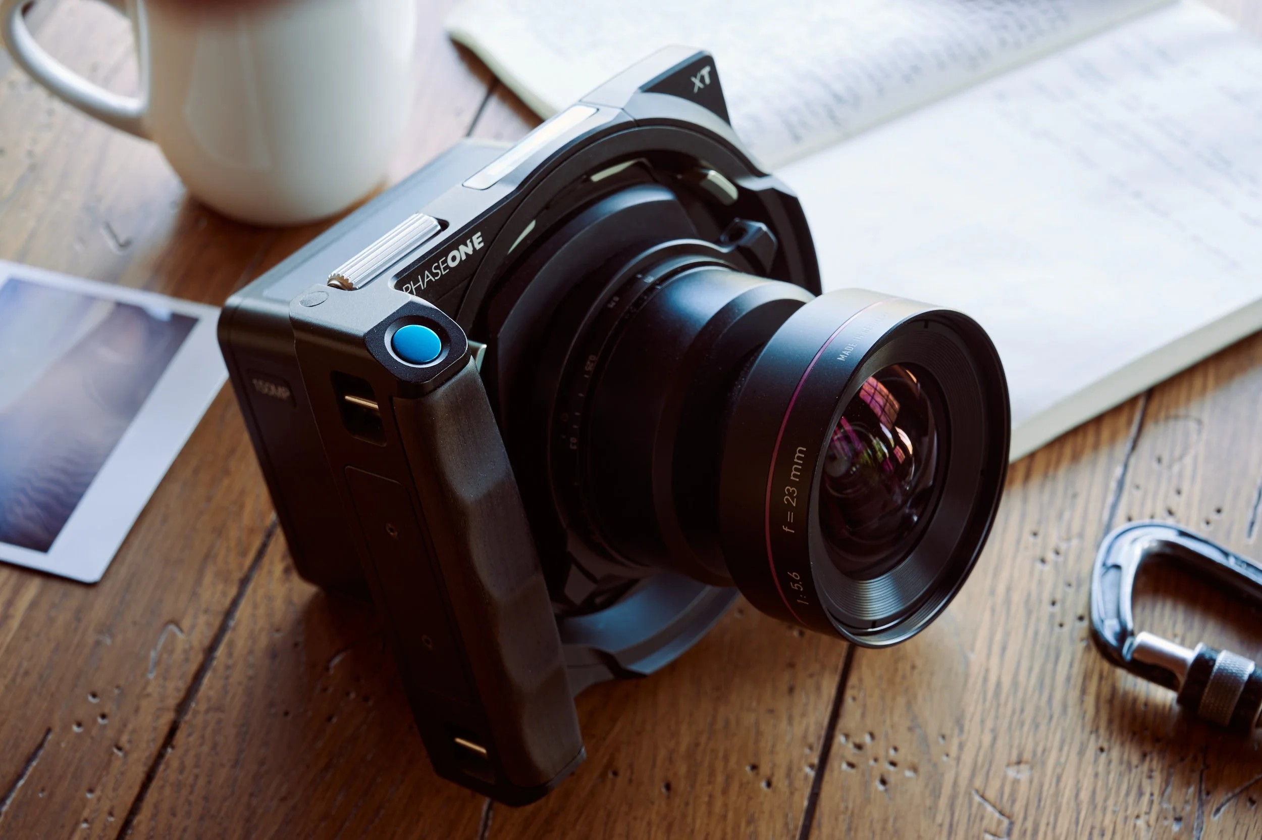 A professional camera with a large lens on a wooden table, alongside a cup, a printed photo, a magazine, and a tripod clamp.