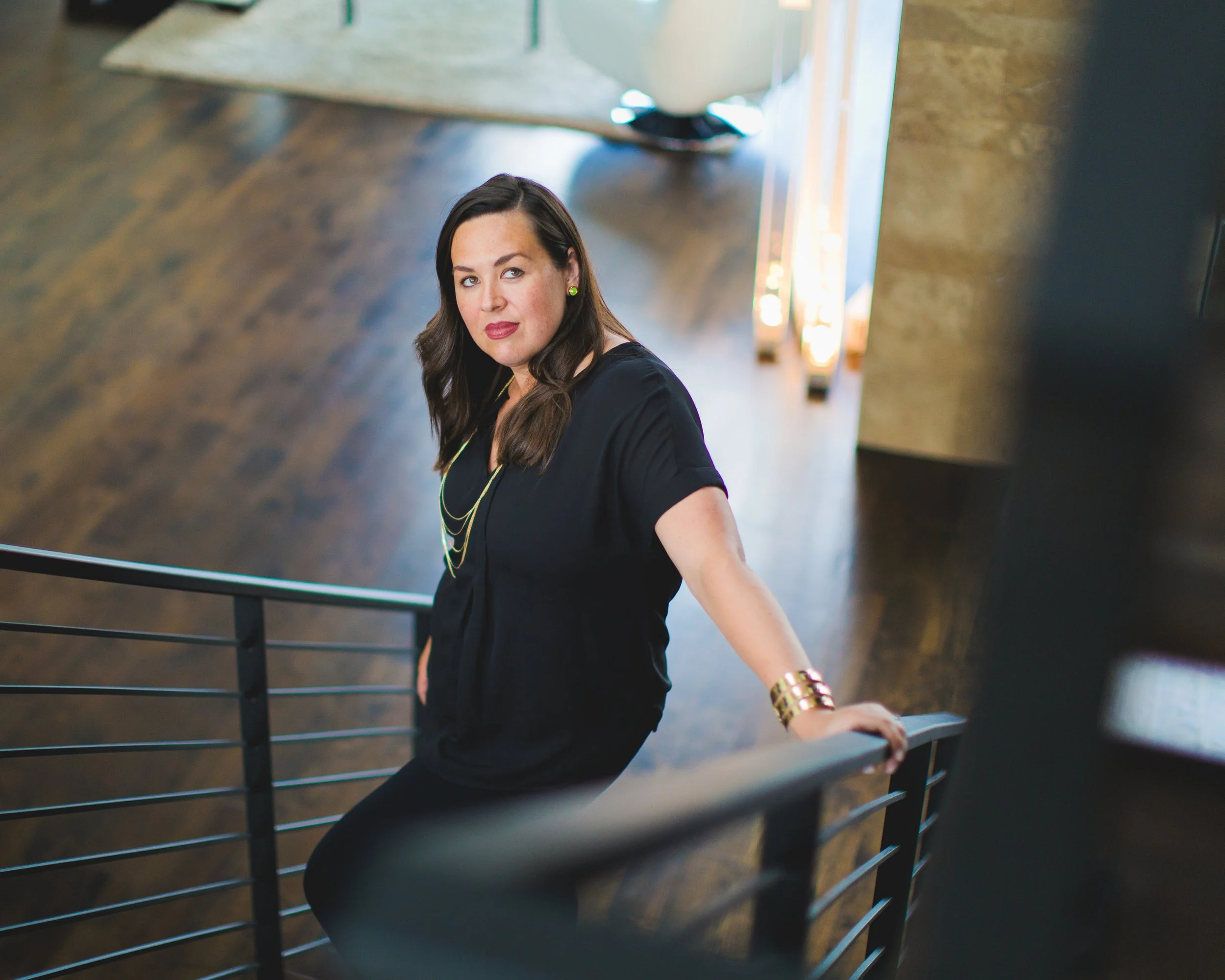 A woman with long brown hair, wearing a black top, gold jewelry, and green earrings, is sitting on a staircase railing and looking at the camera in an indoor setting with wooden flooring and modern decor.