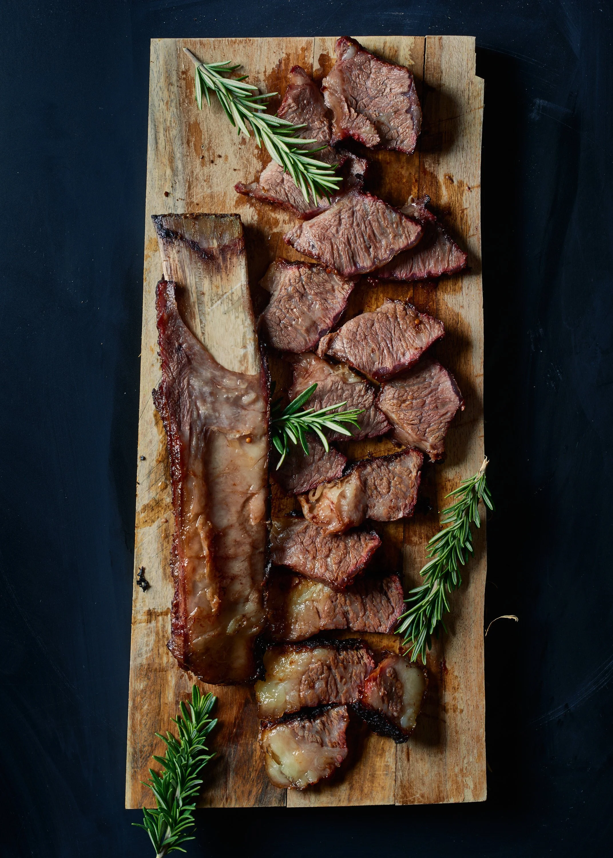 Slices of cooked beef brisket with rosemary sprigs on a wooden cutting board.