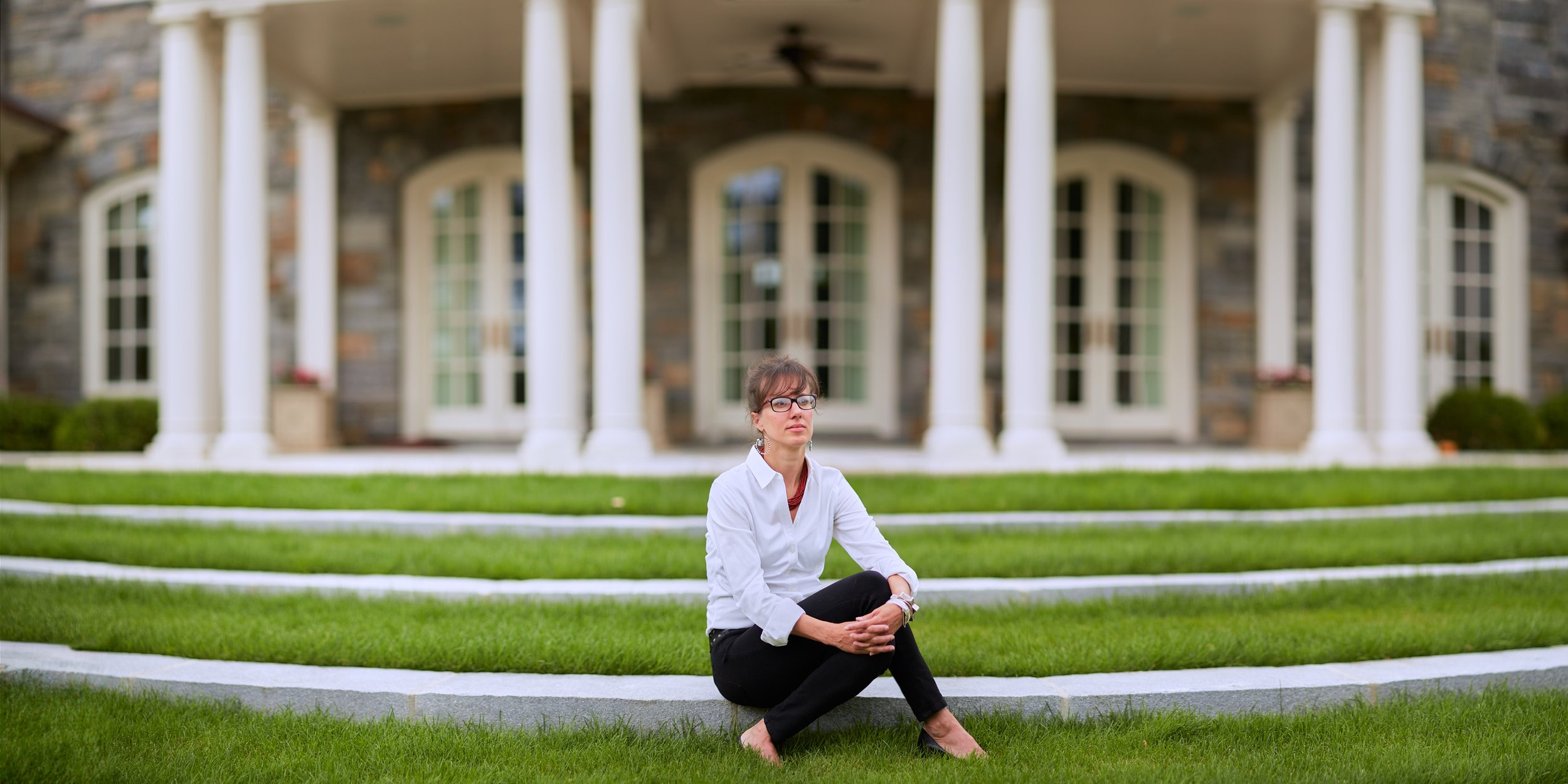 A woman with glasses and a white shirt sitting on grass in front of a large house with columns and multiple windows.