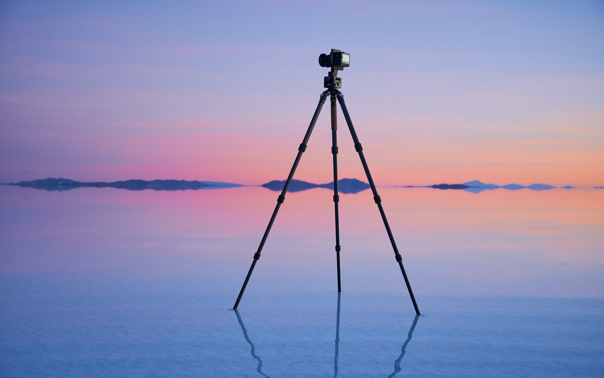 Camera mounted on a tripod standing on a reflective water surface at sunset, with mountains in the background.