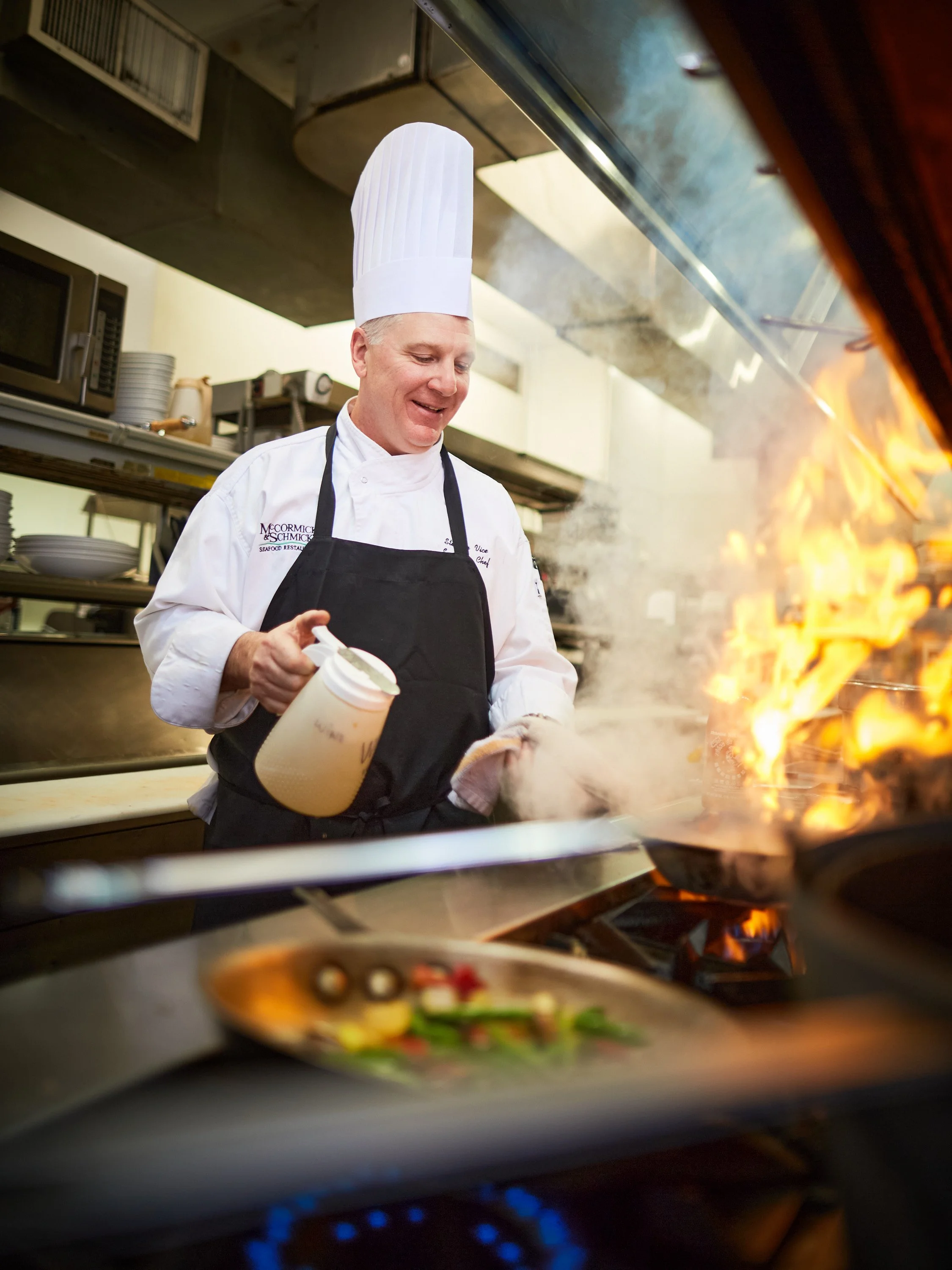 A chef wearing a white chef's hat and black apron cooks on a stove with flames, holding a spray bottle, in a commercial kitchen.