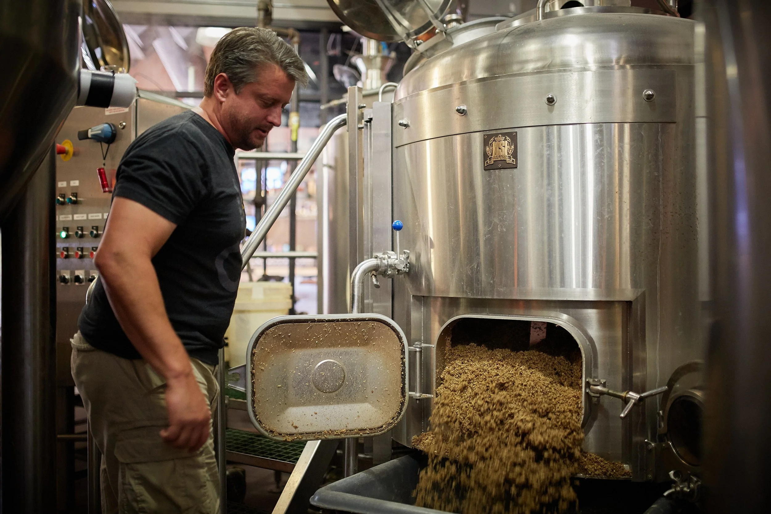 A man in black shirt and khaki shorts standing near a large stainless steel brewing kettle with grains being poured out of it into a container.