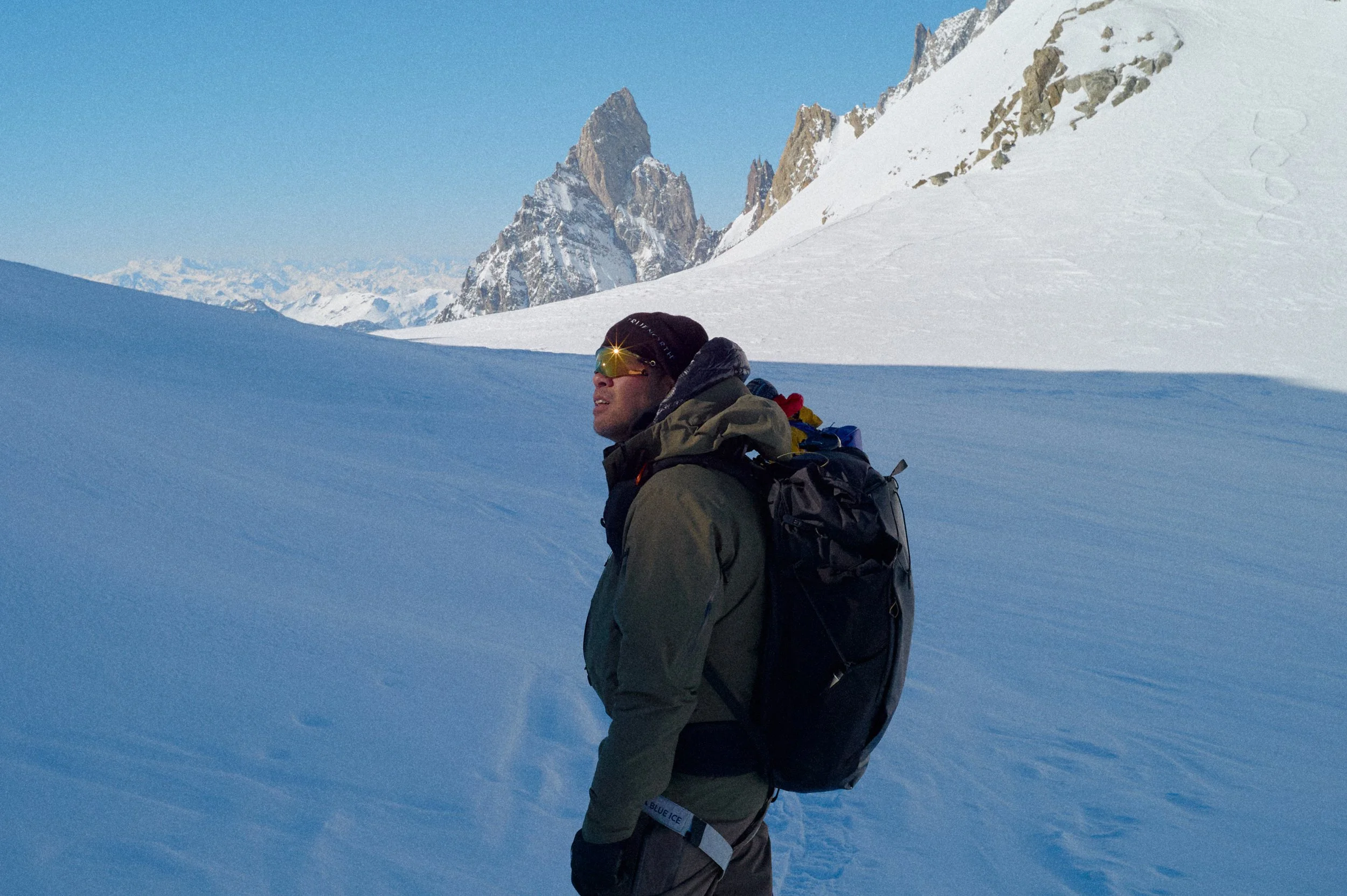 A man stands in a snowy mountain landscape with rocky peaks in the background, wearing winter gear and sunglasses.