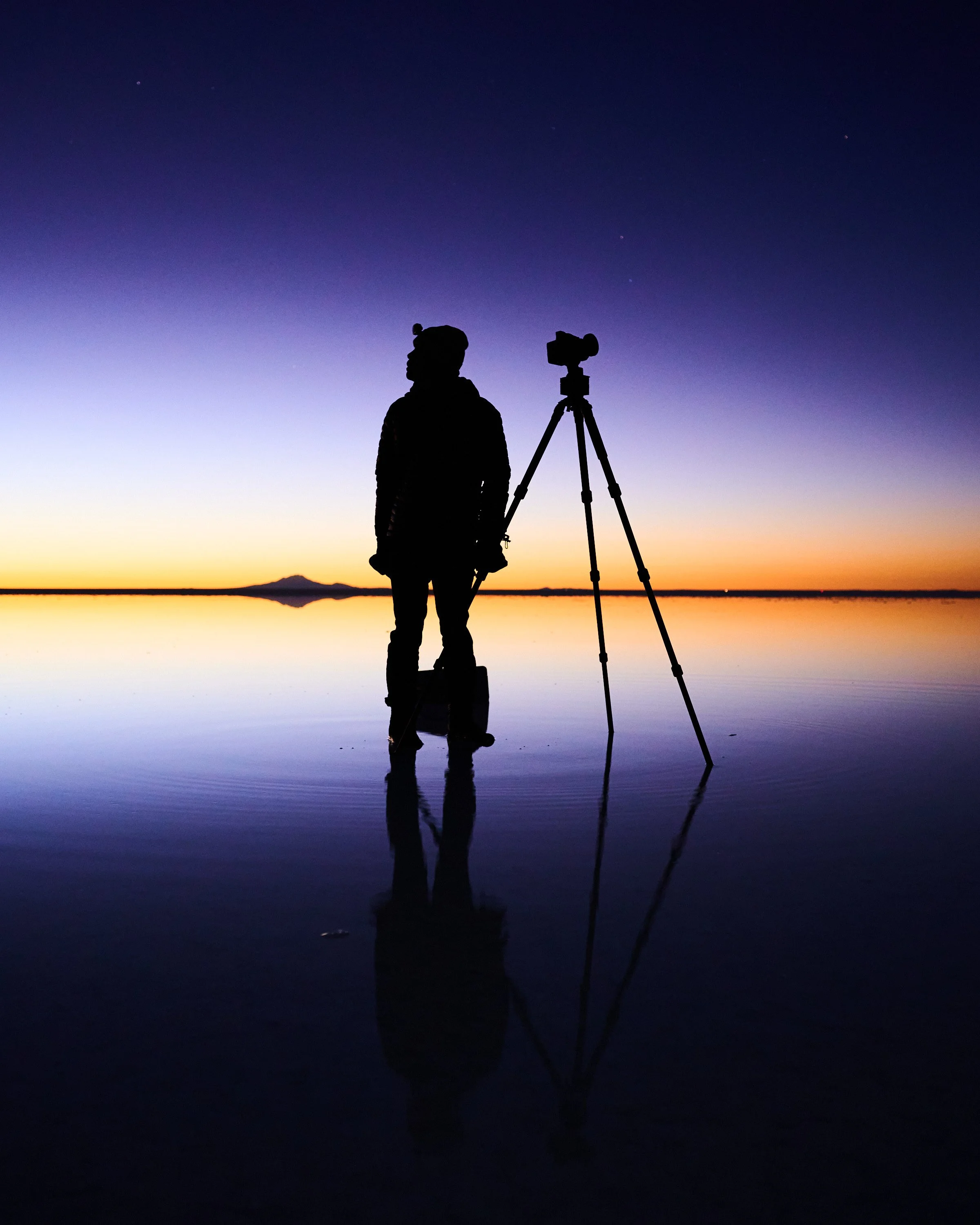 Silhouette of a person with a camera on a tripod standing by a reflective body of water during sunset or sunrise, with a mountain in the distance.