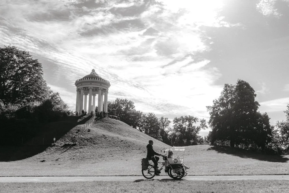 Unter sonnigem Münchner Himmel fährt ein Brautpaar in einer Rickscha durch den ENglischen Garten unter dem Monopterus vorbei.