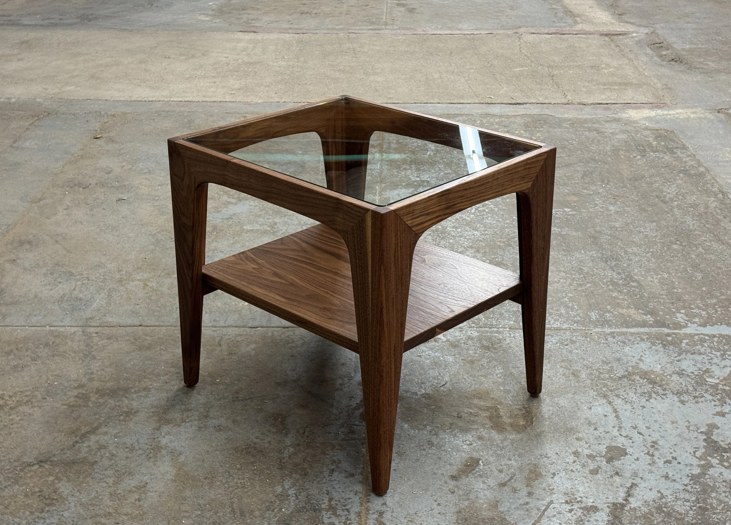 A wooden side table with a glass top and lower wooden shelf, standing on a concrete floor