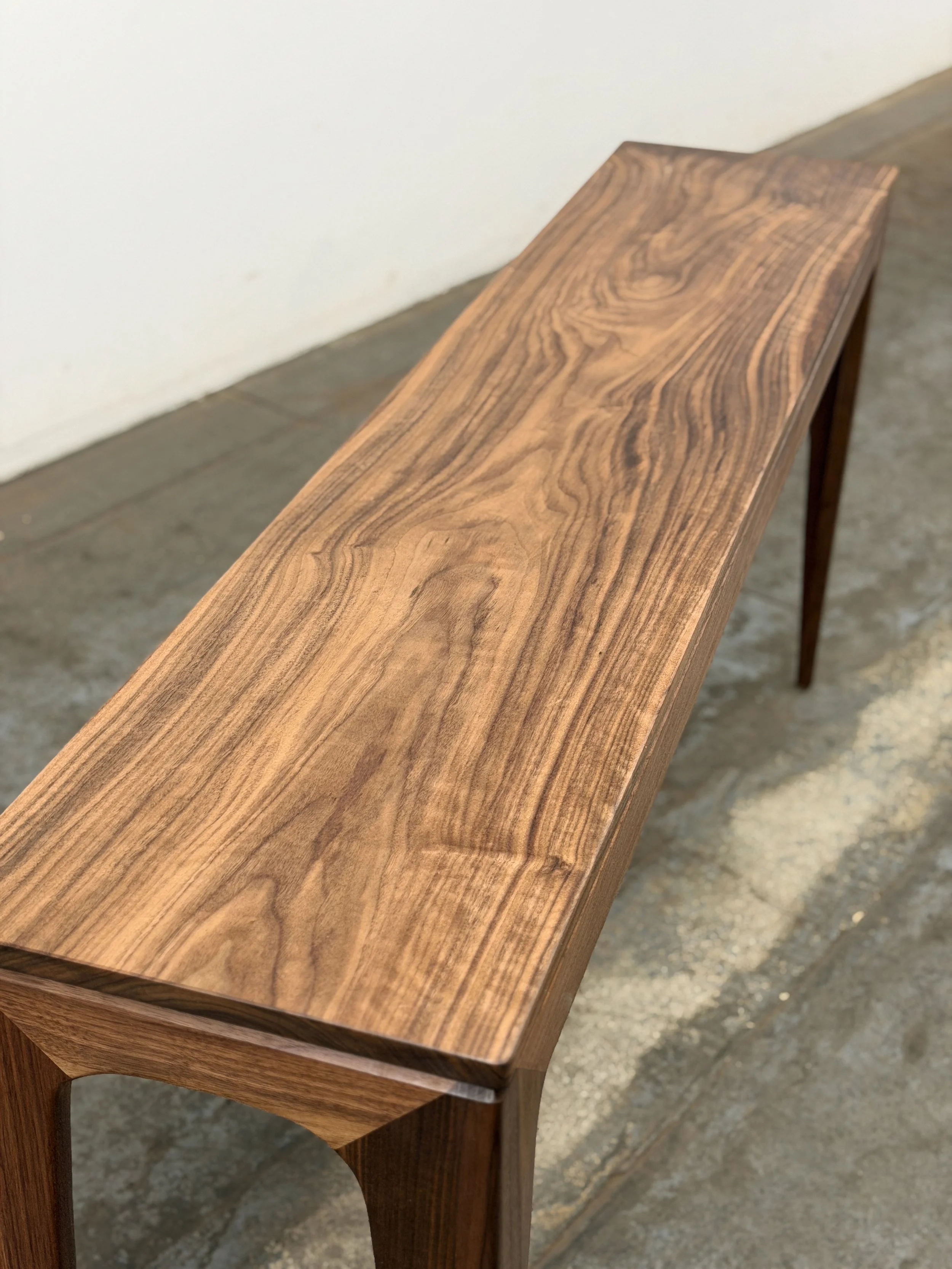 A long wooden table with a natural wood grain finish, supported by four matching wooden legs, placed against a white wall on a concrete floor.