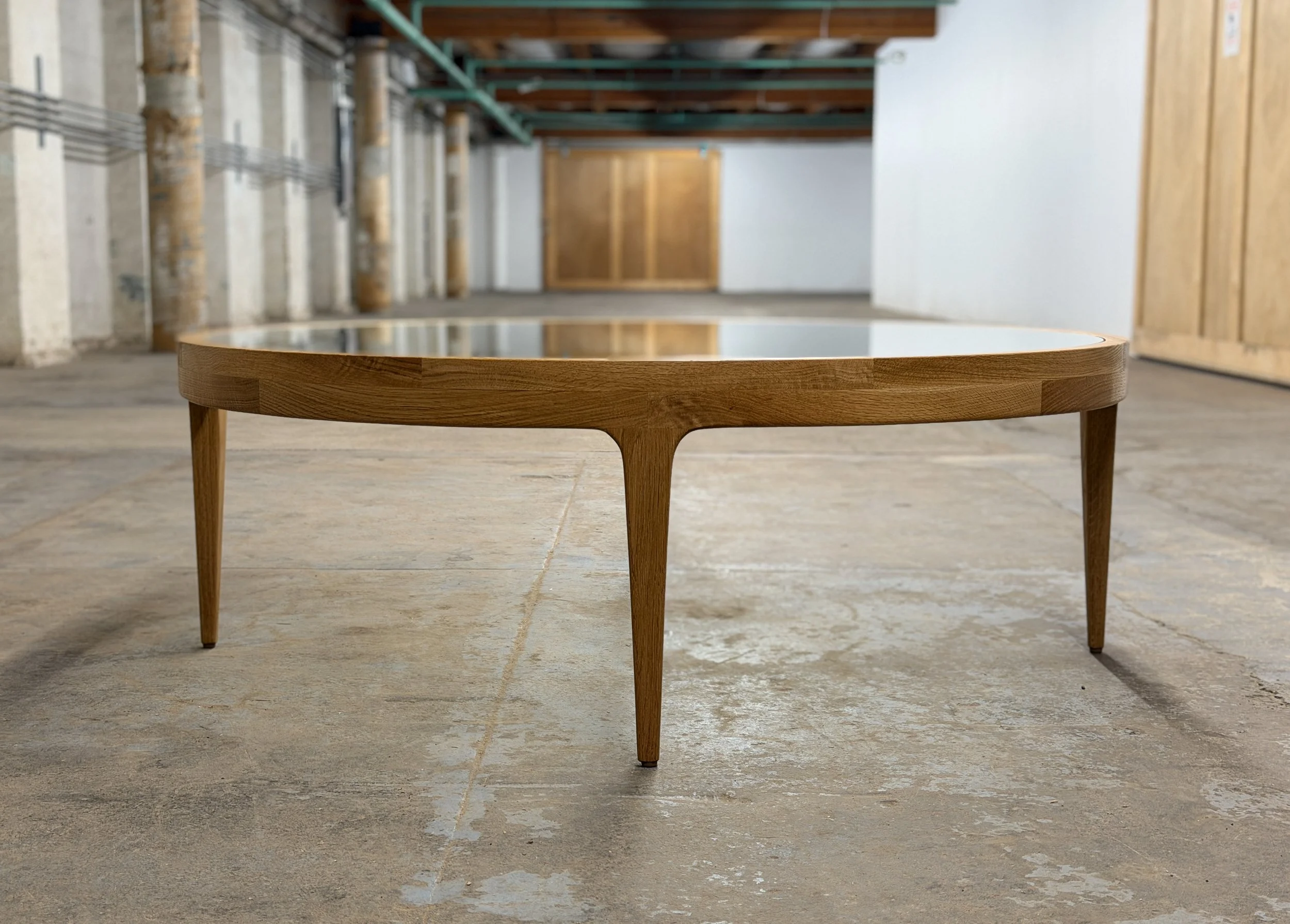 A modern wooden coffee table with a glass top, placed on a concrete floor in an industrial-style room with exposed pipes and wooden sliding doors.