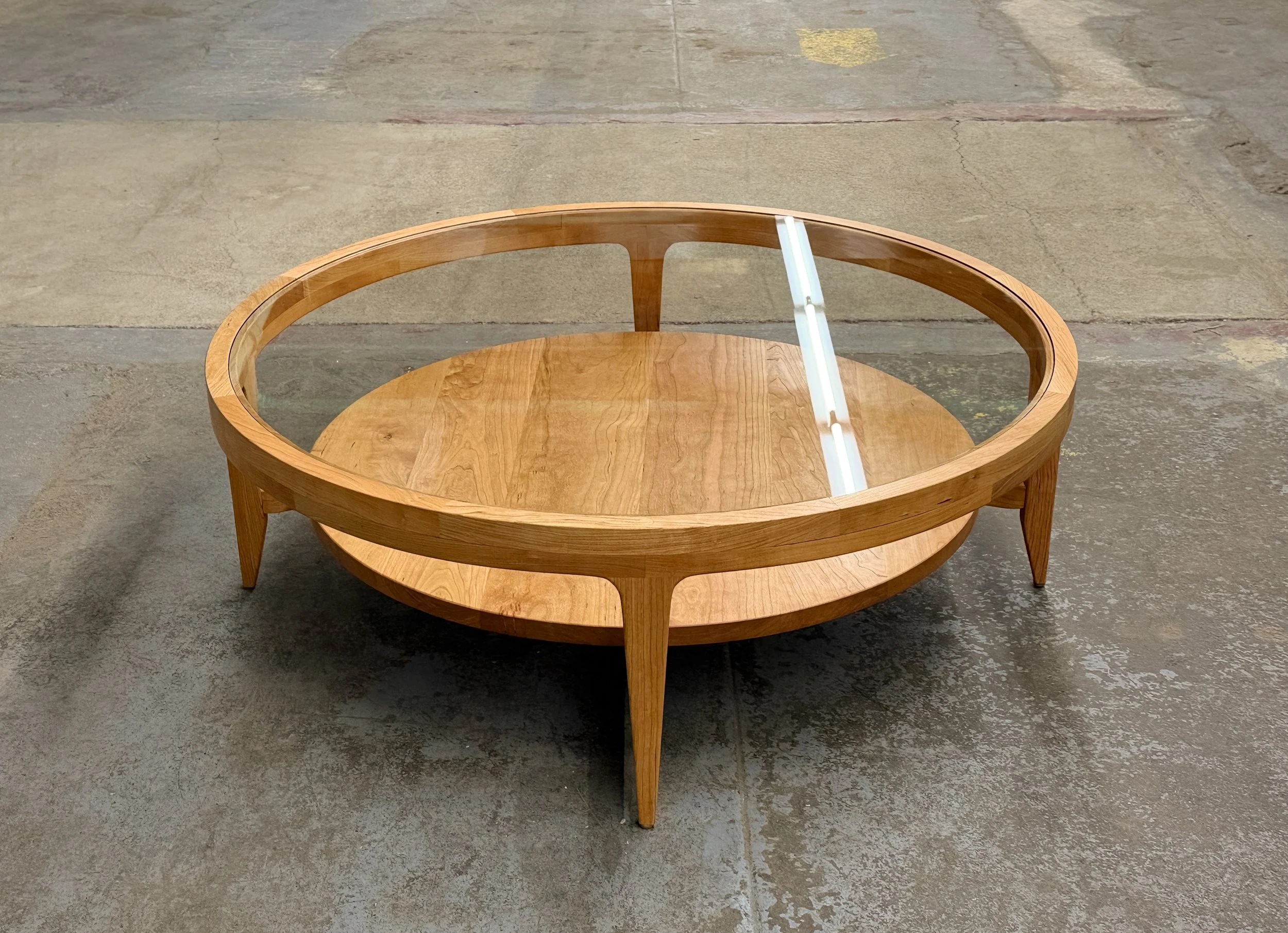 Round wooden and glass coffee table with a lower wooden shelf, situated on a concrete floor.