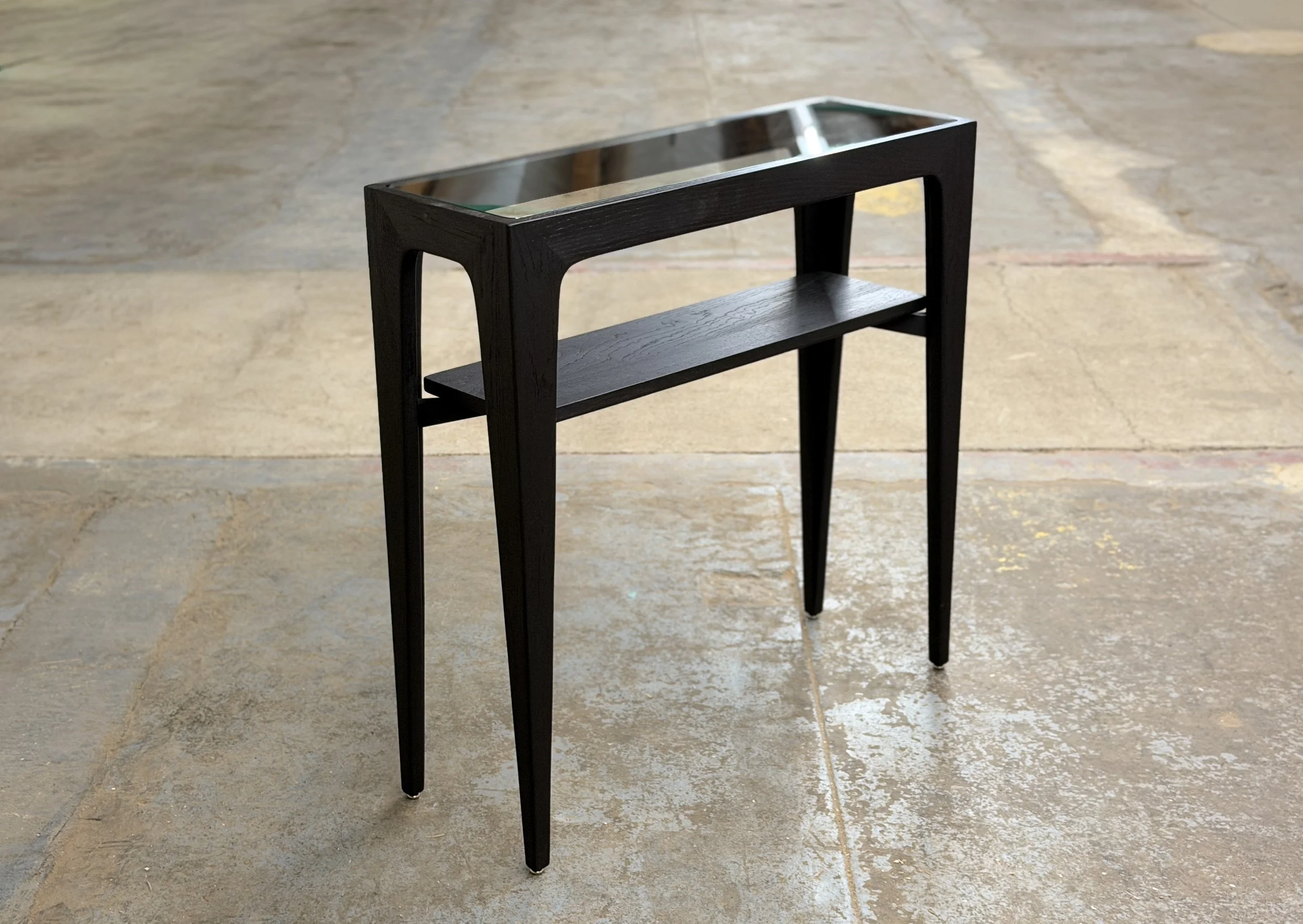 A small black wooden side table with a glass top and a lower shelf, standing on a tiled floor in a room with natural light.
