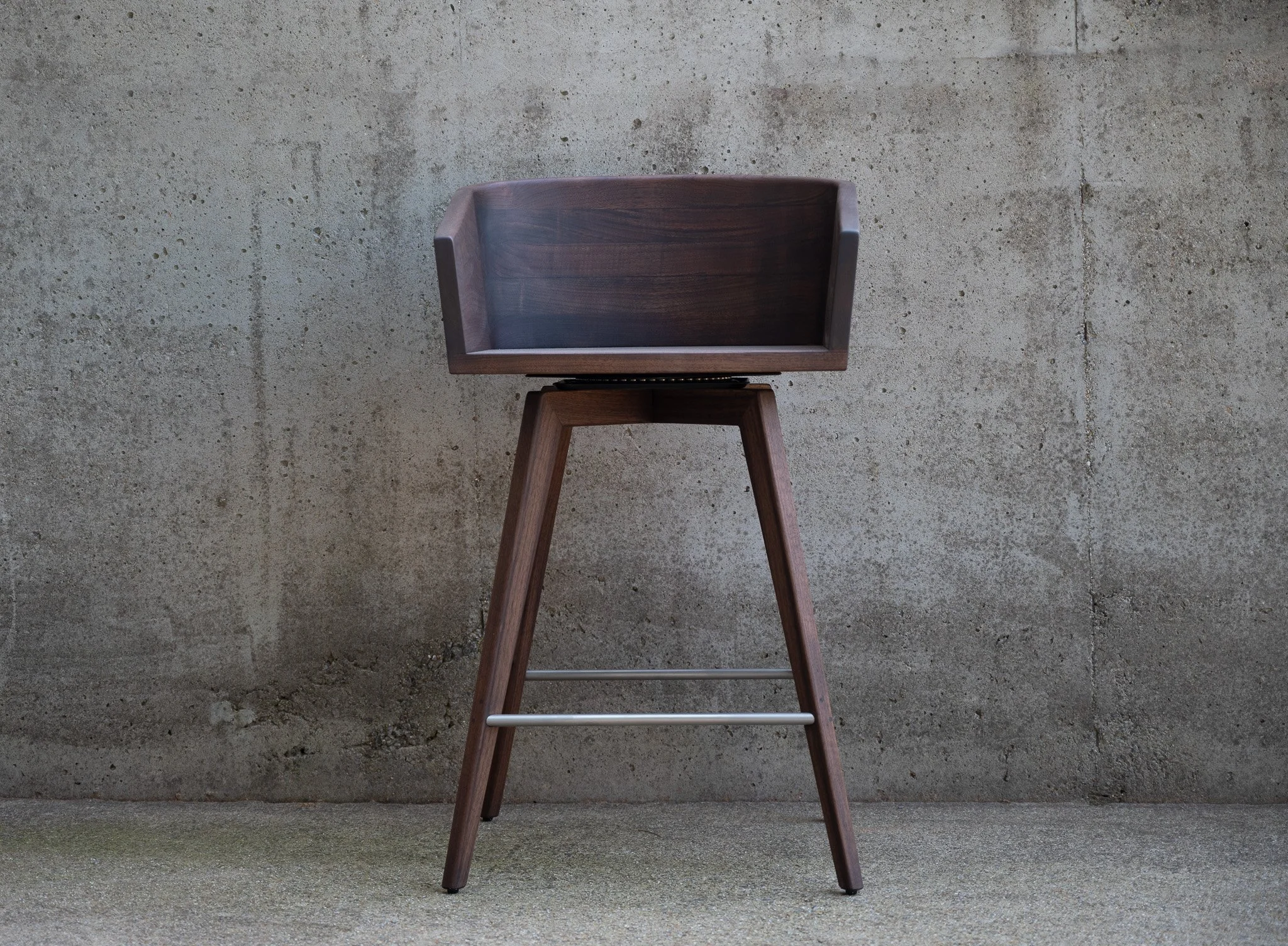 A wooden chair with a high back and angled legs, positioned against a textured concrete wall.