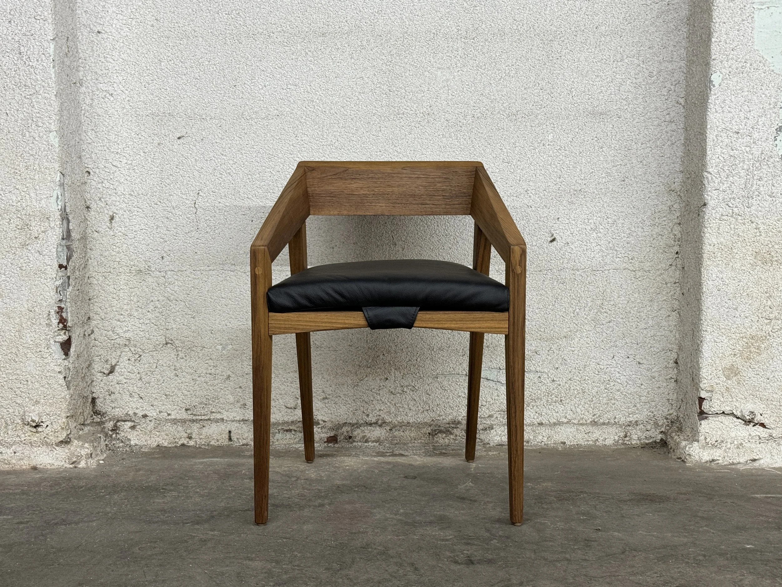A wooden chair with a black cushioned seat is placed against a textured white wall on a concrete floor.