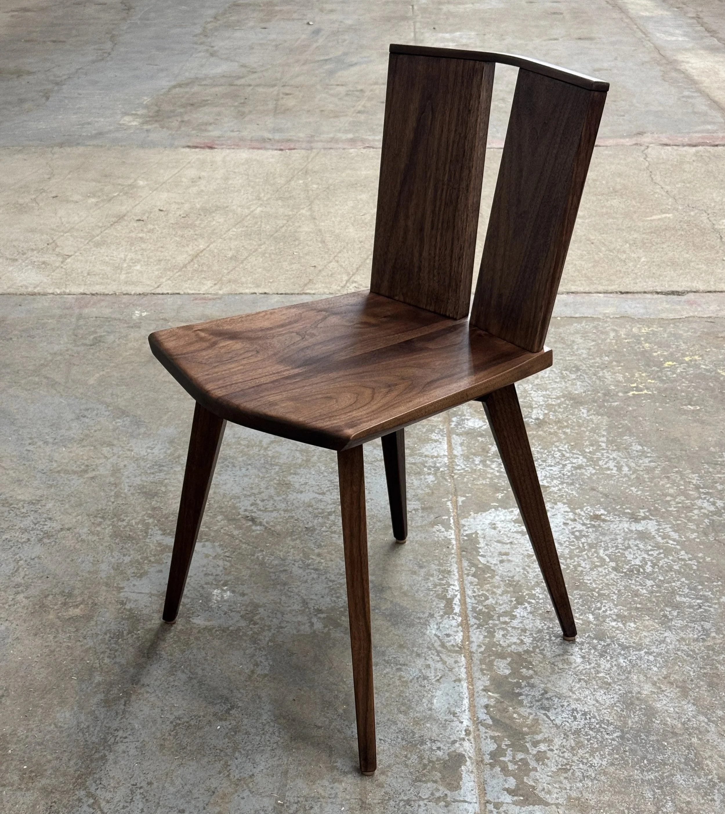 A wooden chair with angled backrest and four tapered legs placed on a concrete floor.