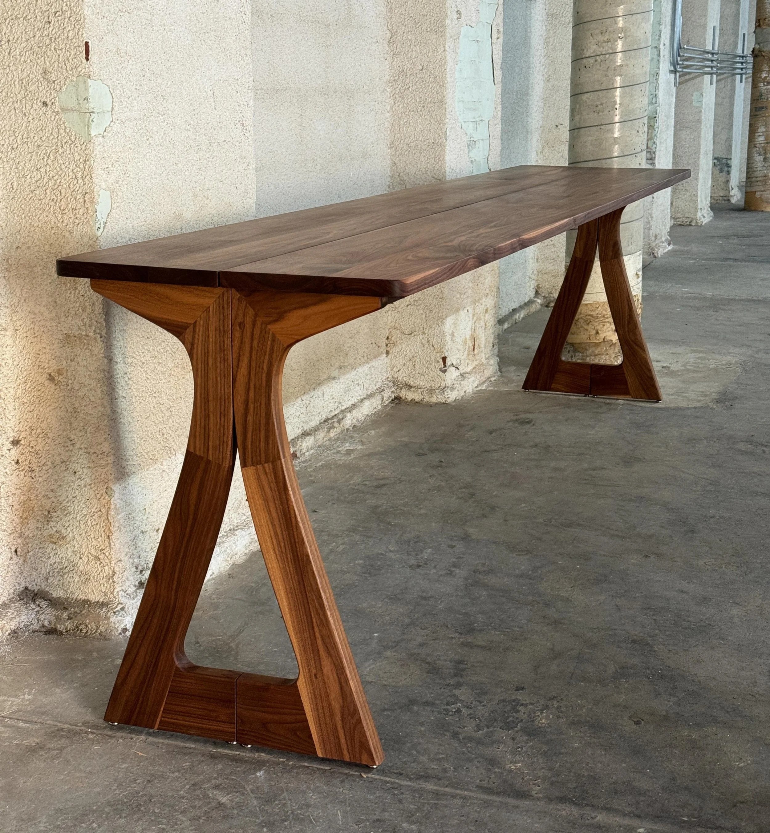 Wooden console table with curved pedestal legs in an industrial space with concrete floor and textured walls.