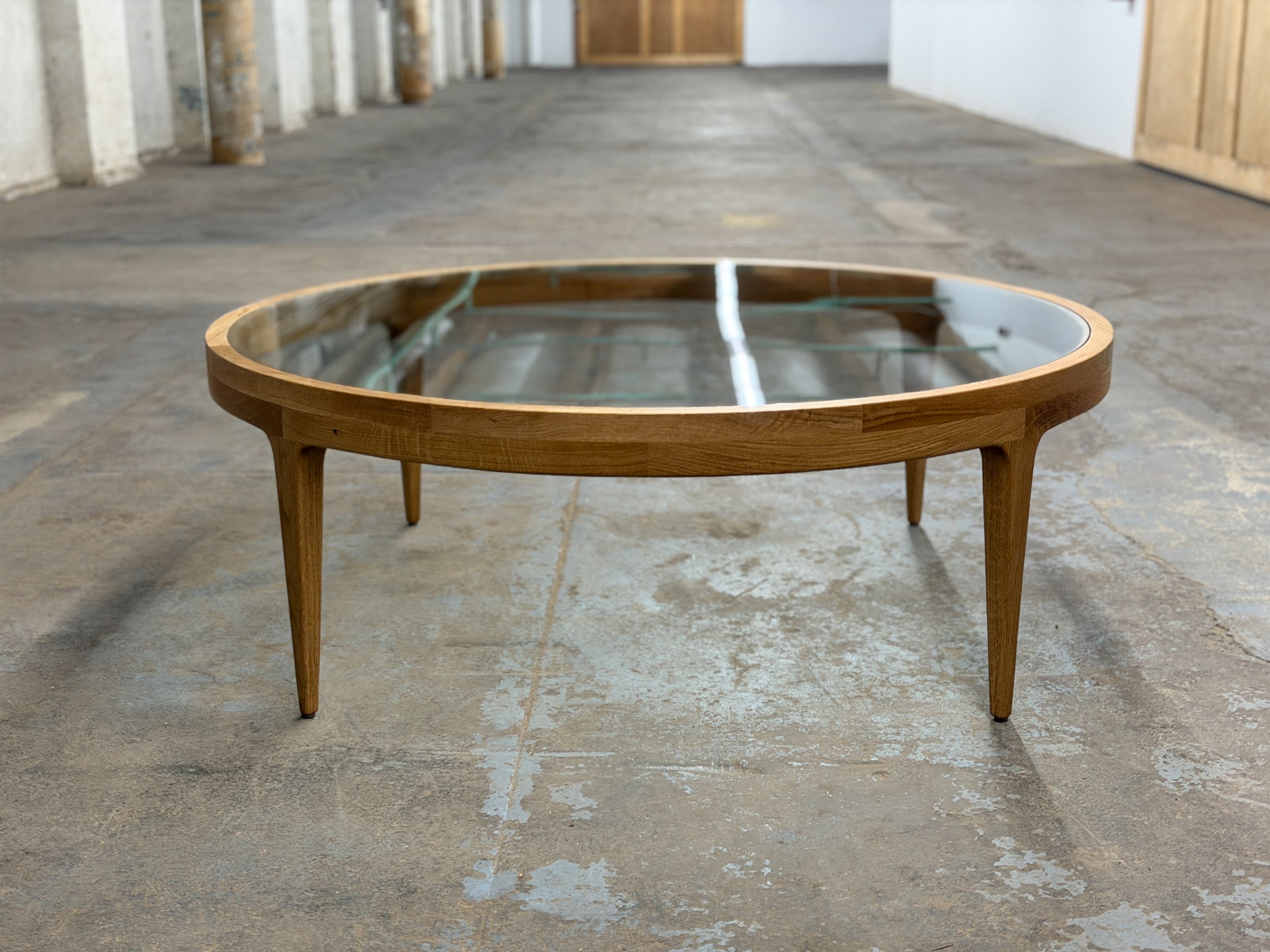 A mid-century modern wooden coffee table with a glass top in an unfinished room with a concrete floor.