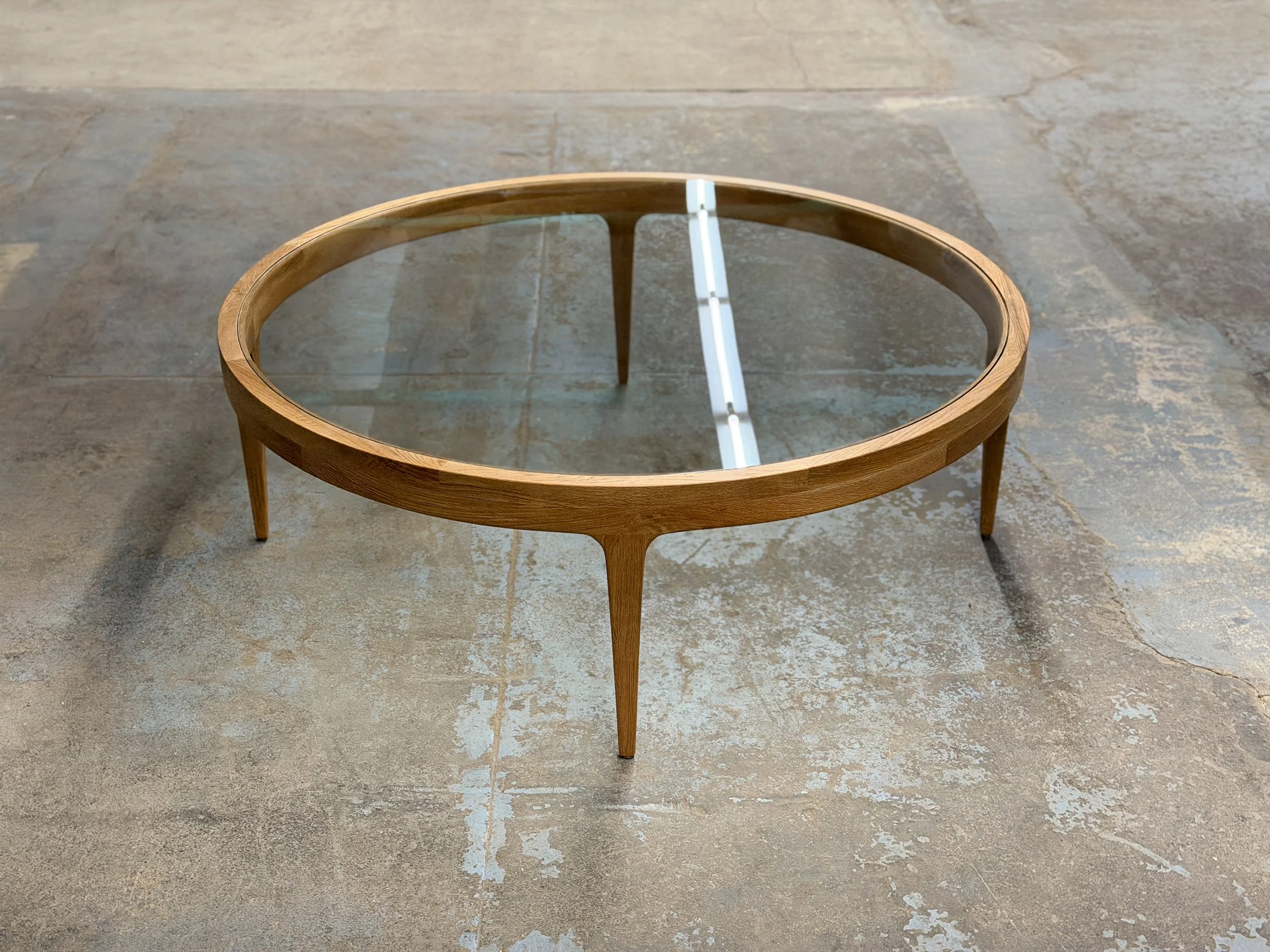 A round wooden coffee table with a glass top, showing a reflection of a light fixture above, placed on a concrete floor with visible paint and wear.