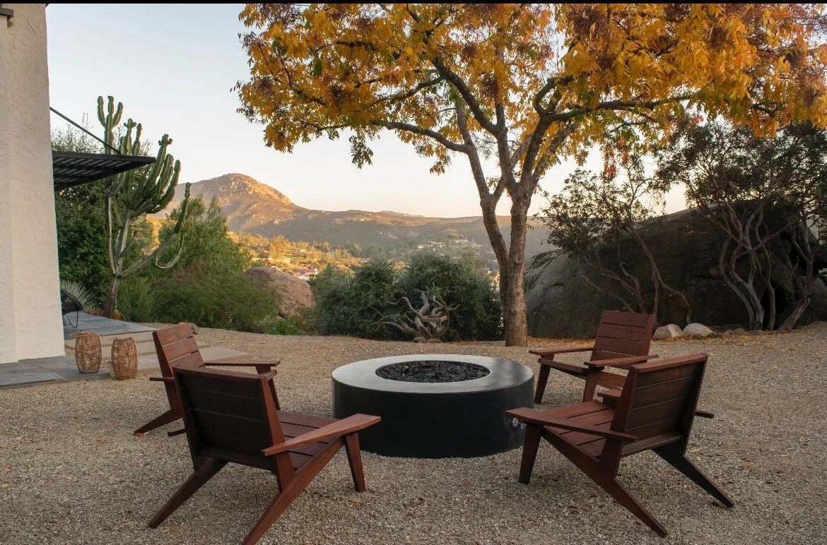 Outdoor patio with three wooden chairs arranged around a circular fire pit in a natural landscape with trees and mountains in the background during sunset.