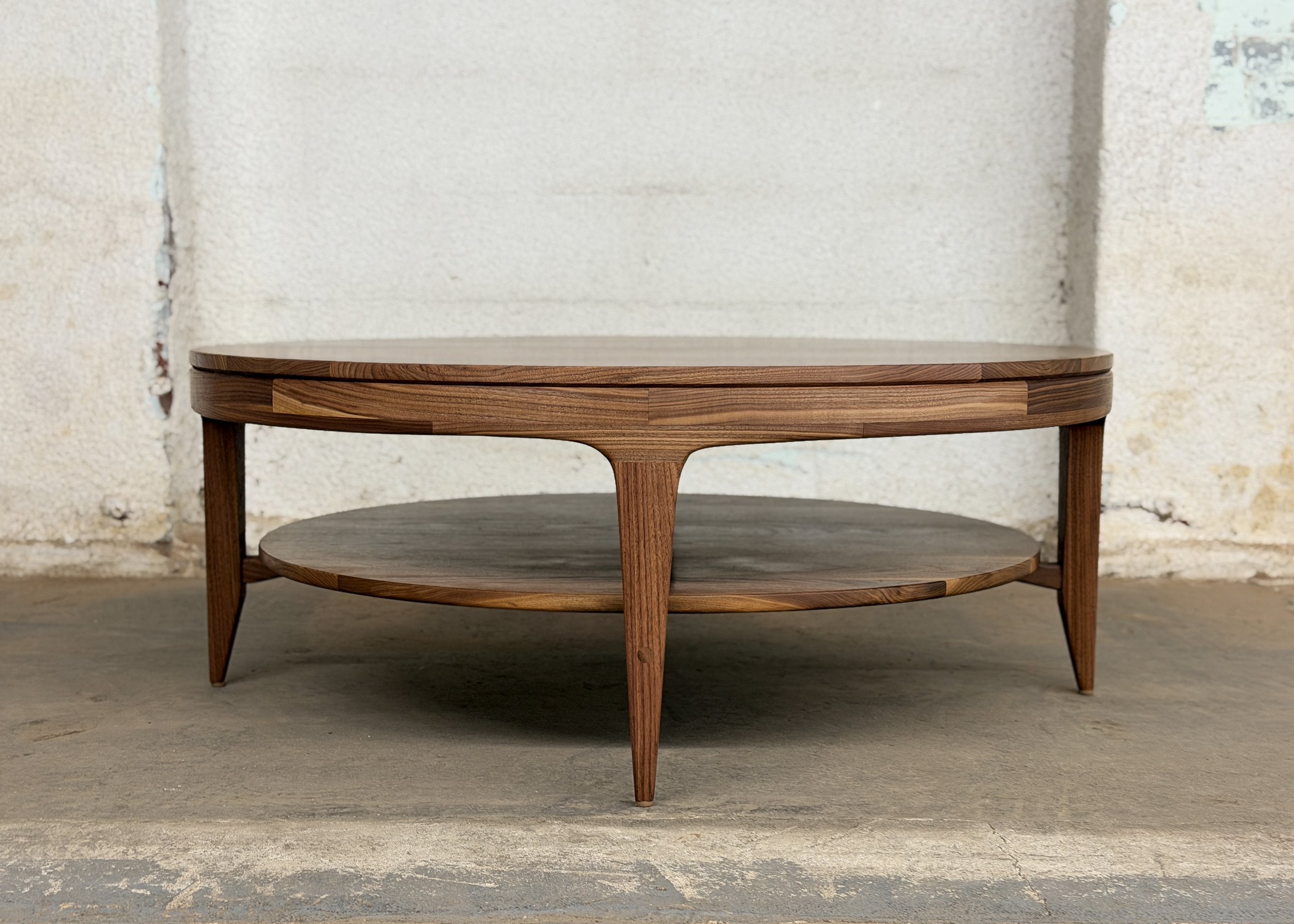 A wooden two-tier coffee table with a round top and a lower shelf, set against a white brick wall.