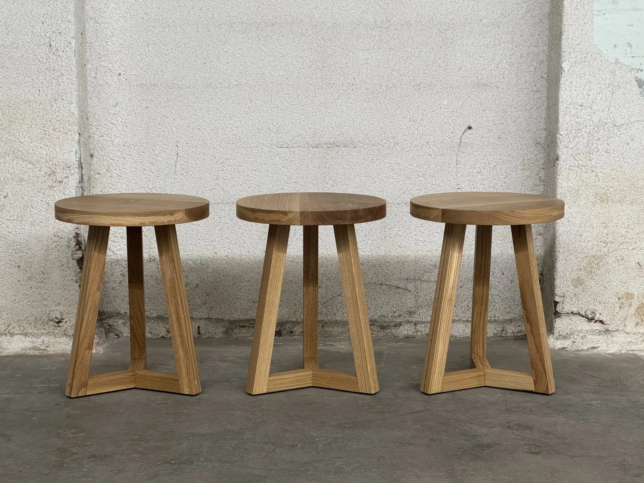 Three wooden stools with round seats and angled legs, lined up against a textured off-white wall.