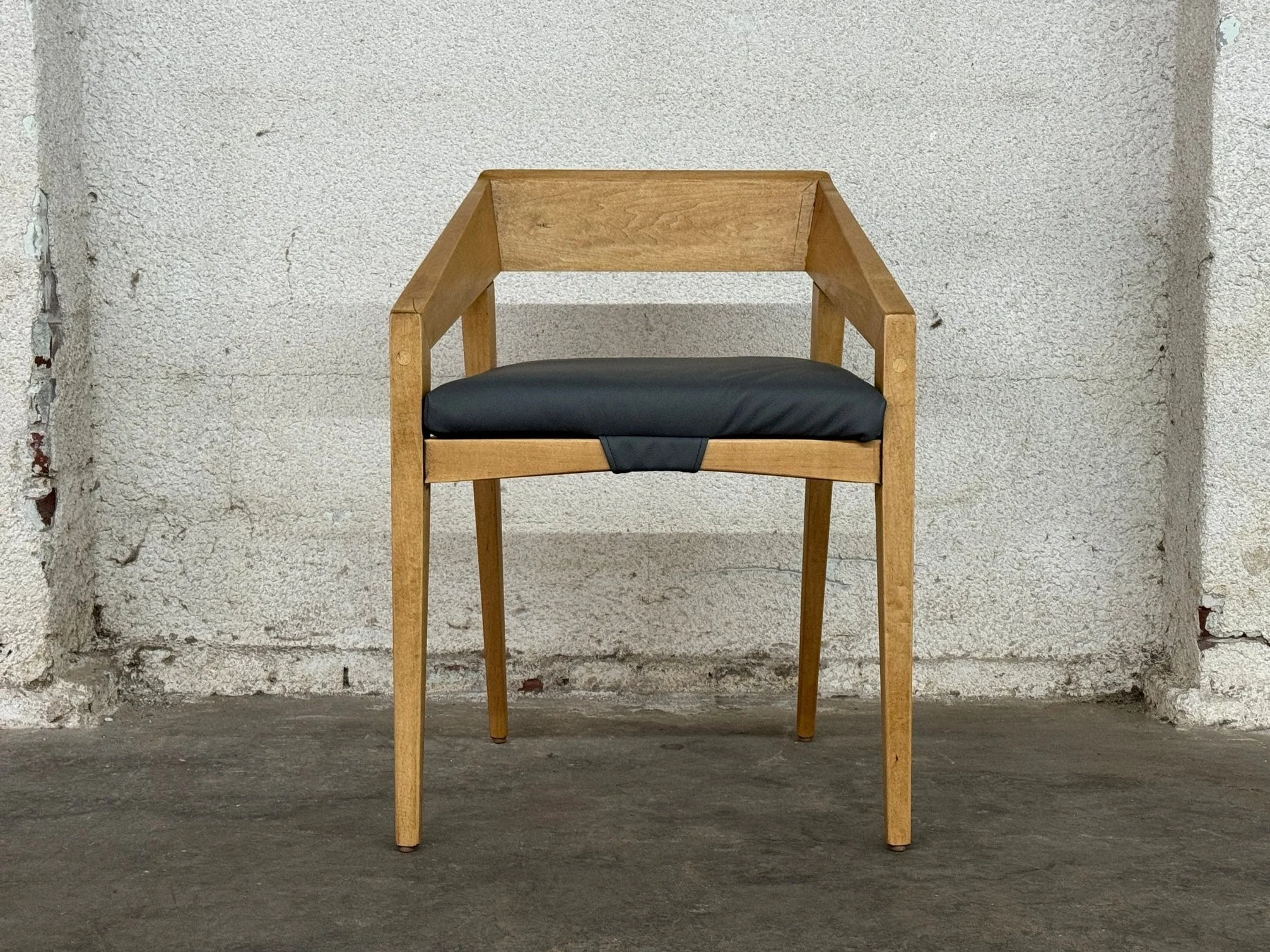 A wooden chair with a black cushioned seat, positioned against a textured white wall and concrete floor.
