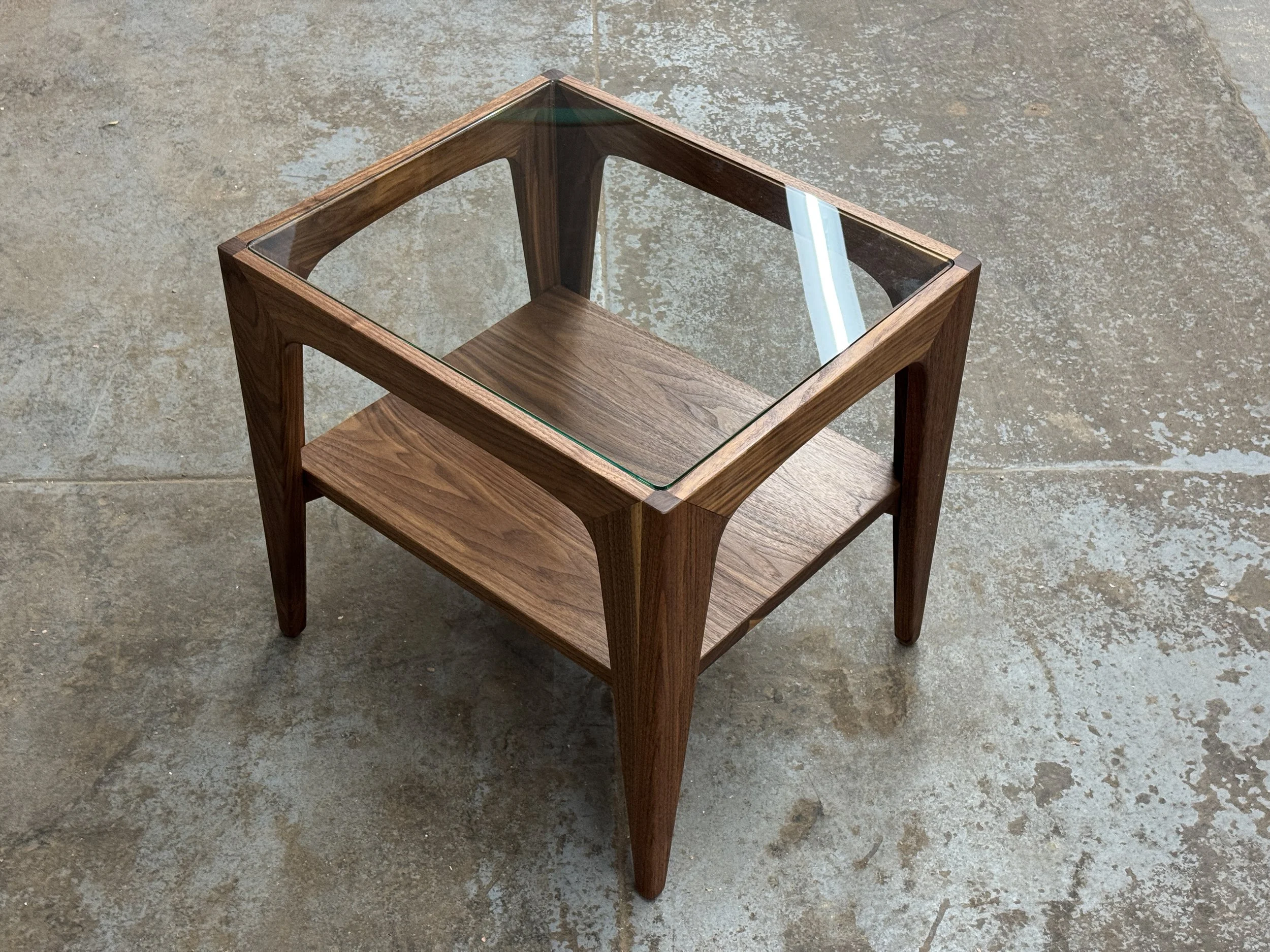 A modern wooden side table with a glass top, standing on a concrete floor.
