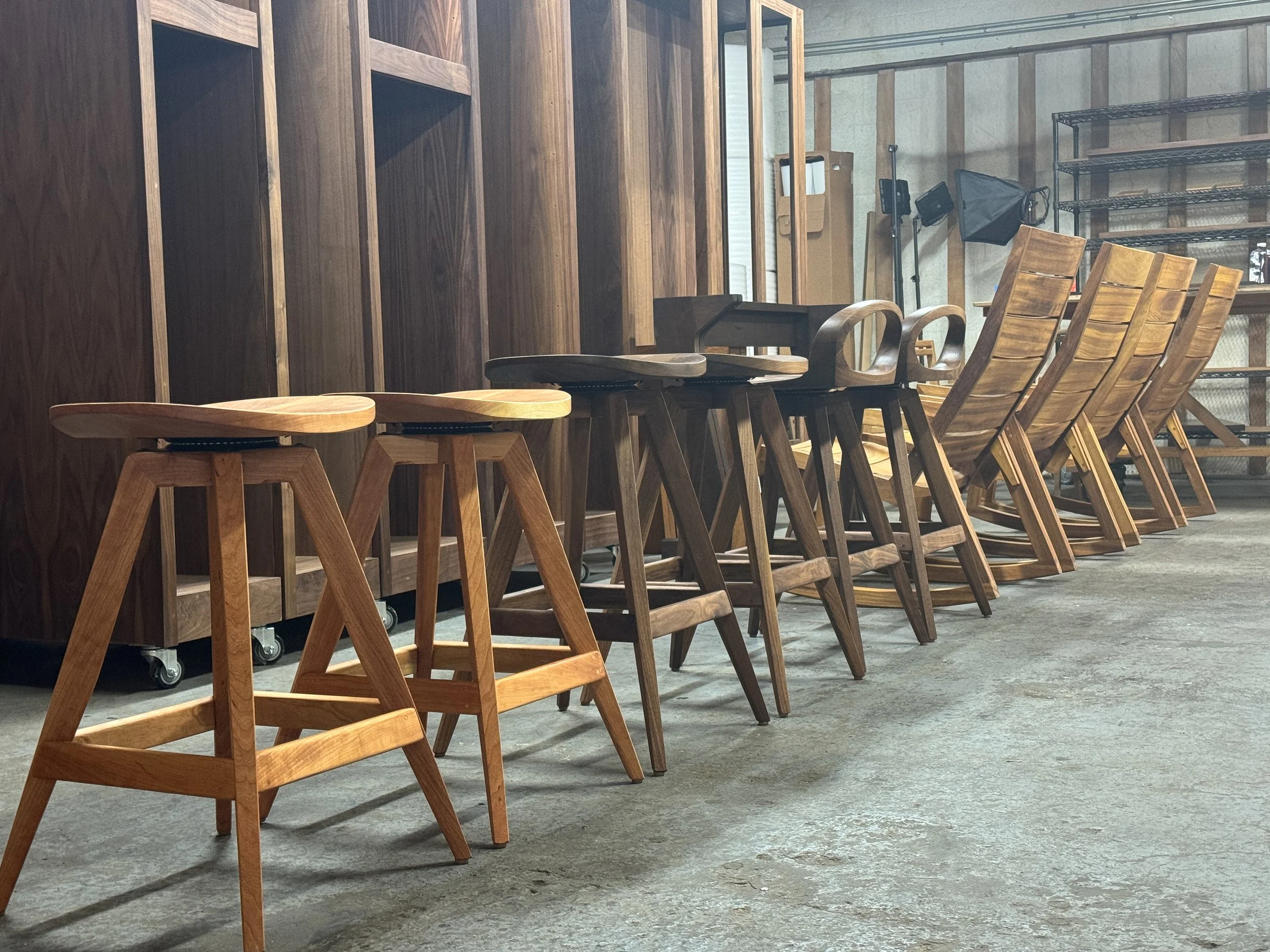 A row of wooden chairs and stools arranged in an industrial-style room with concrete floor and metal shelving.