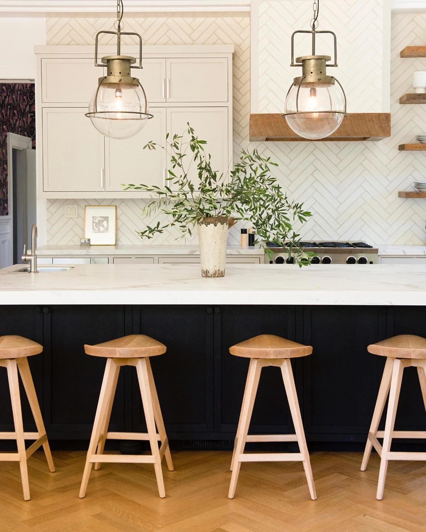 Kitchen with white cabinets, a white marble island, four wooden stools, pendant lights, and a large plant in a beige vase.