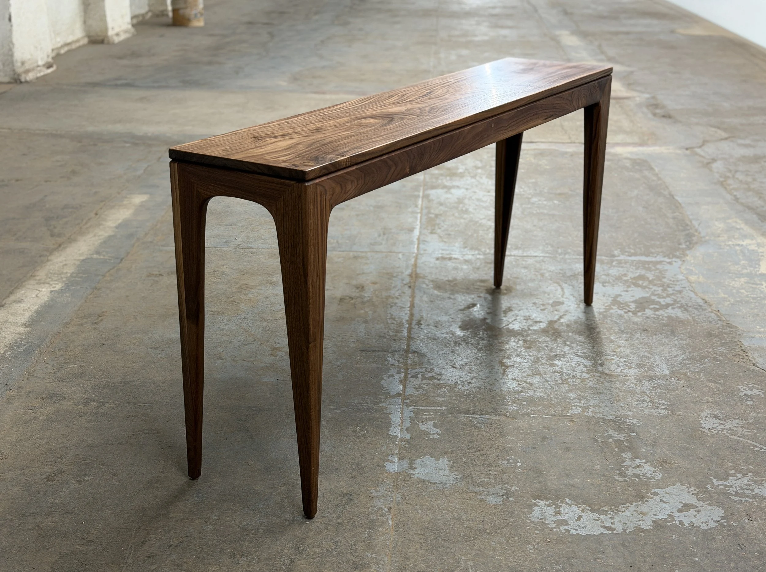 A wooden console table with tapered legs in a rustic setting with concrete flooring and white walls.