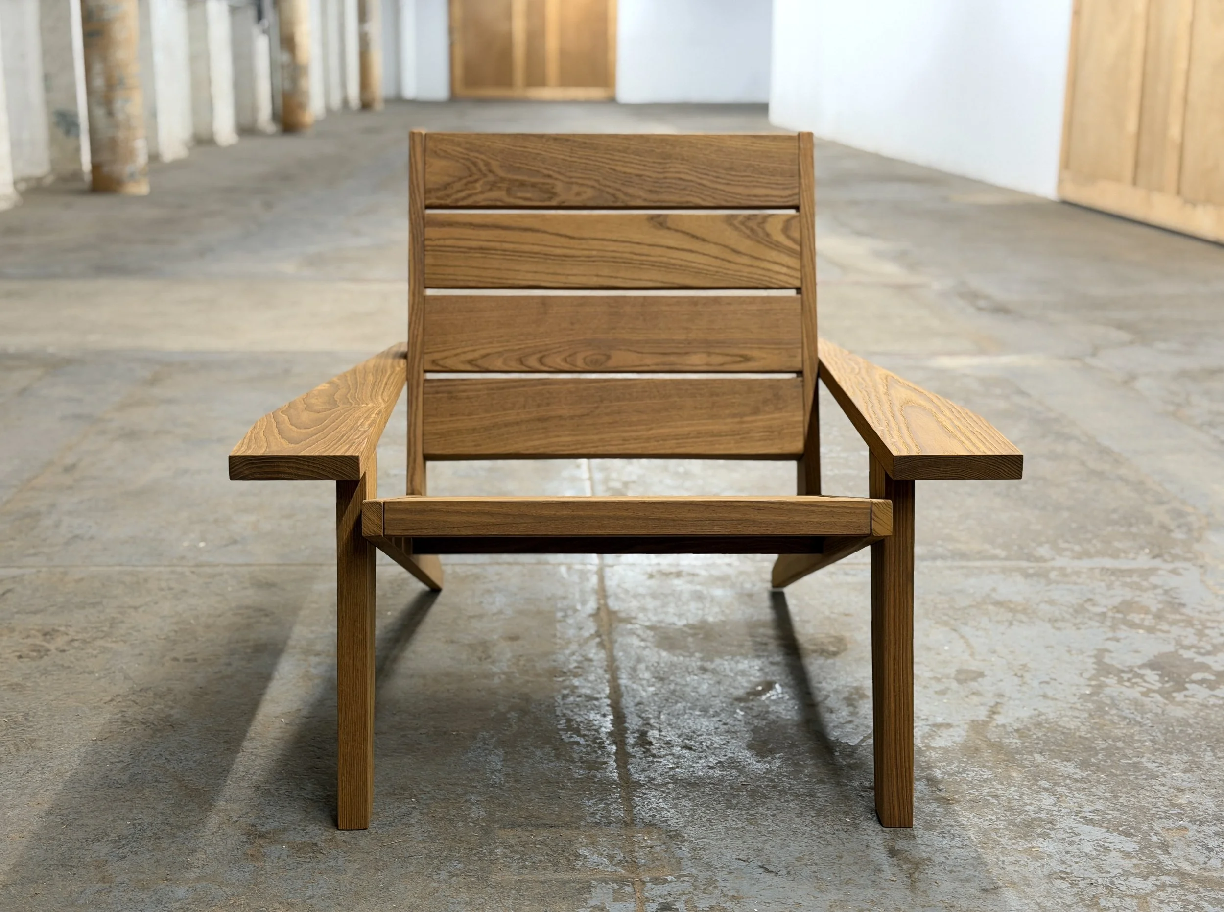 front view of a wooden outdoor chair with armrests, on a concrete floor, in a rustic indoor setting.