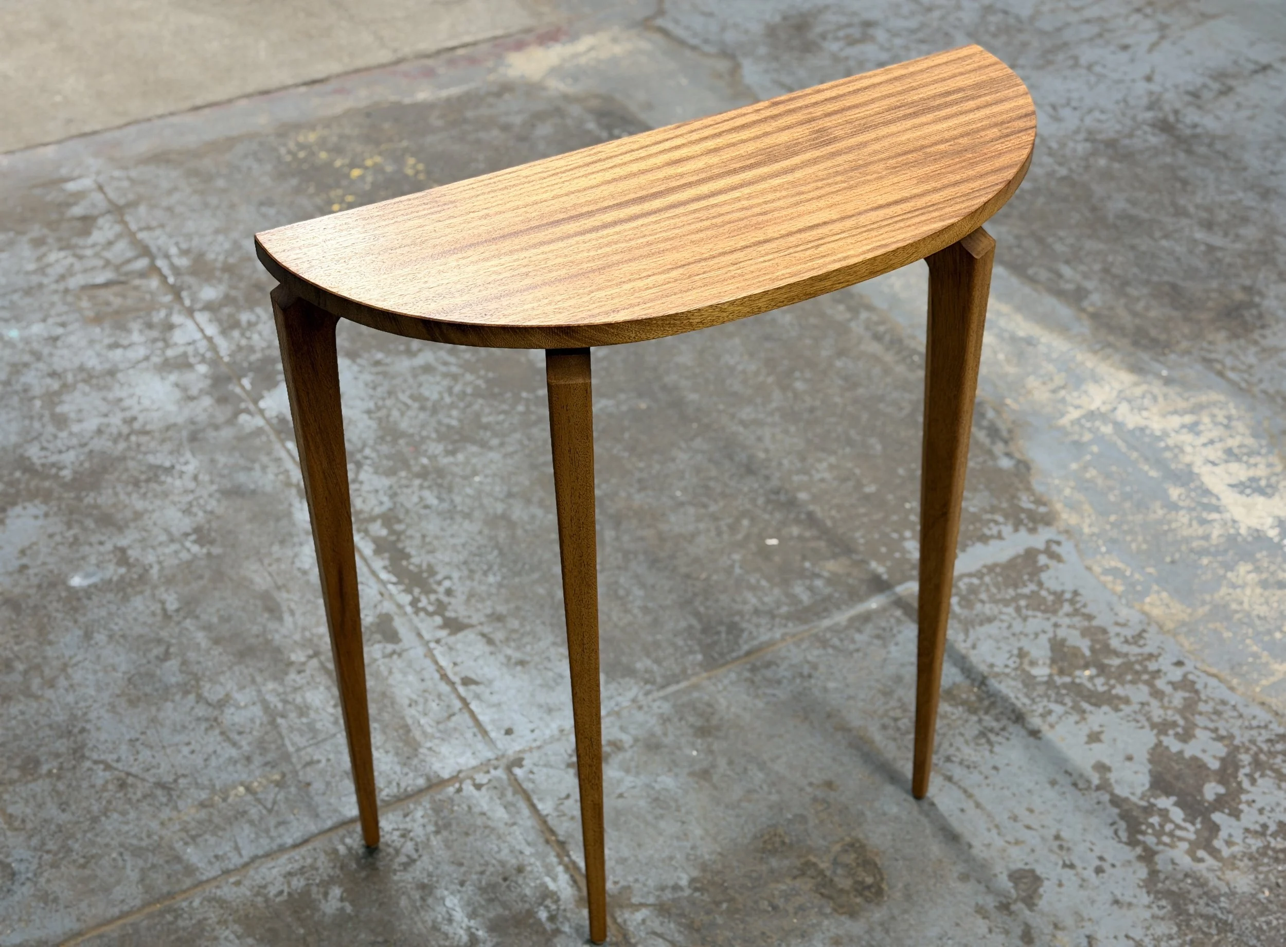 A wooden console table with a curved, semi-circular top and four slender, tapered legs placed on a concrete floor.