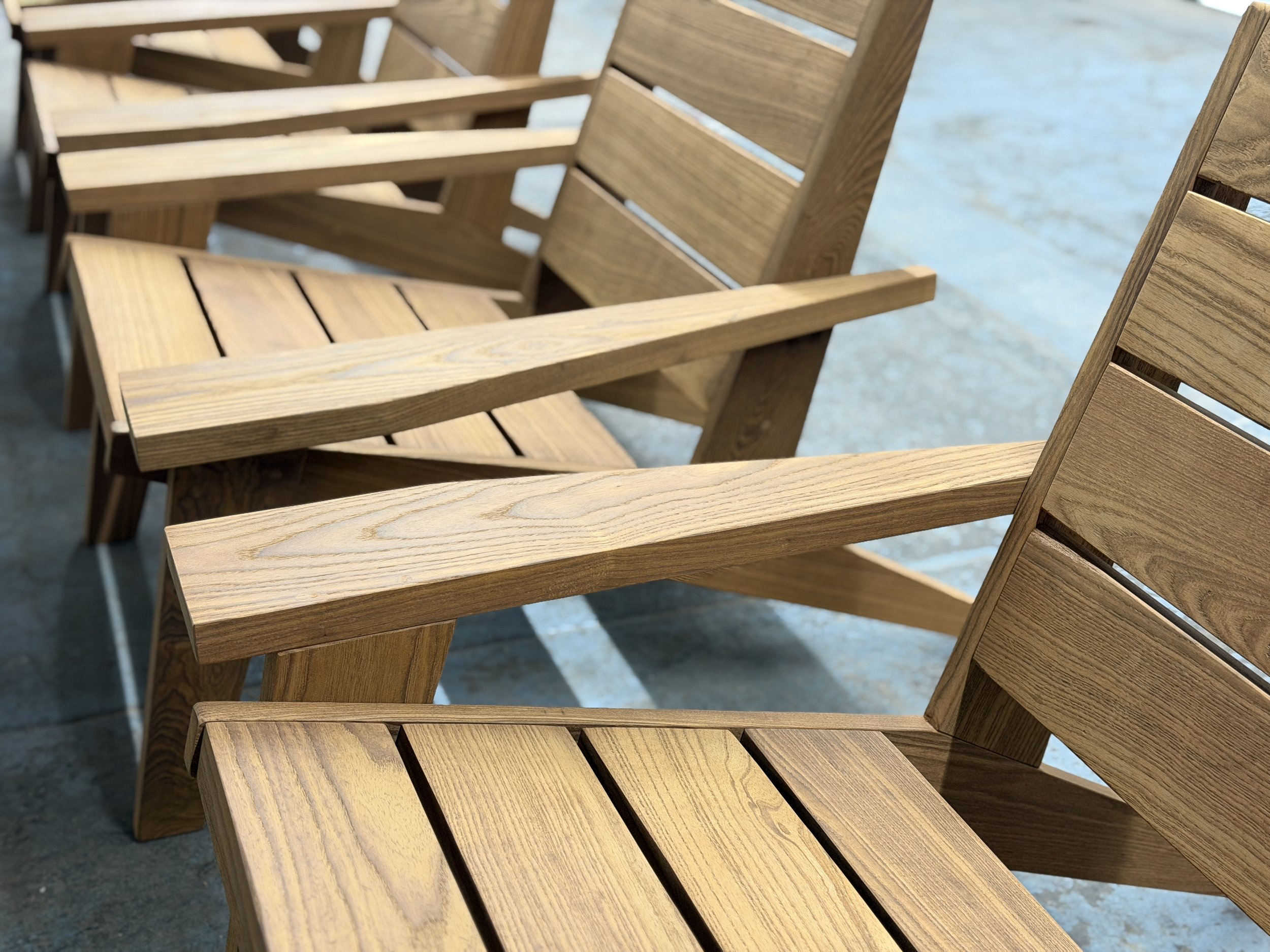 A row of wooden lounge chairs with slatted seats and backrests, arranged on a concrete surface near water.