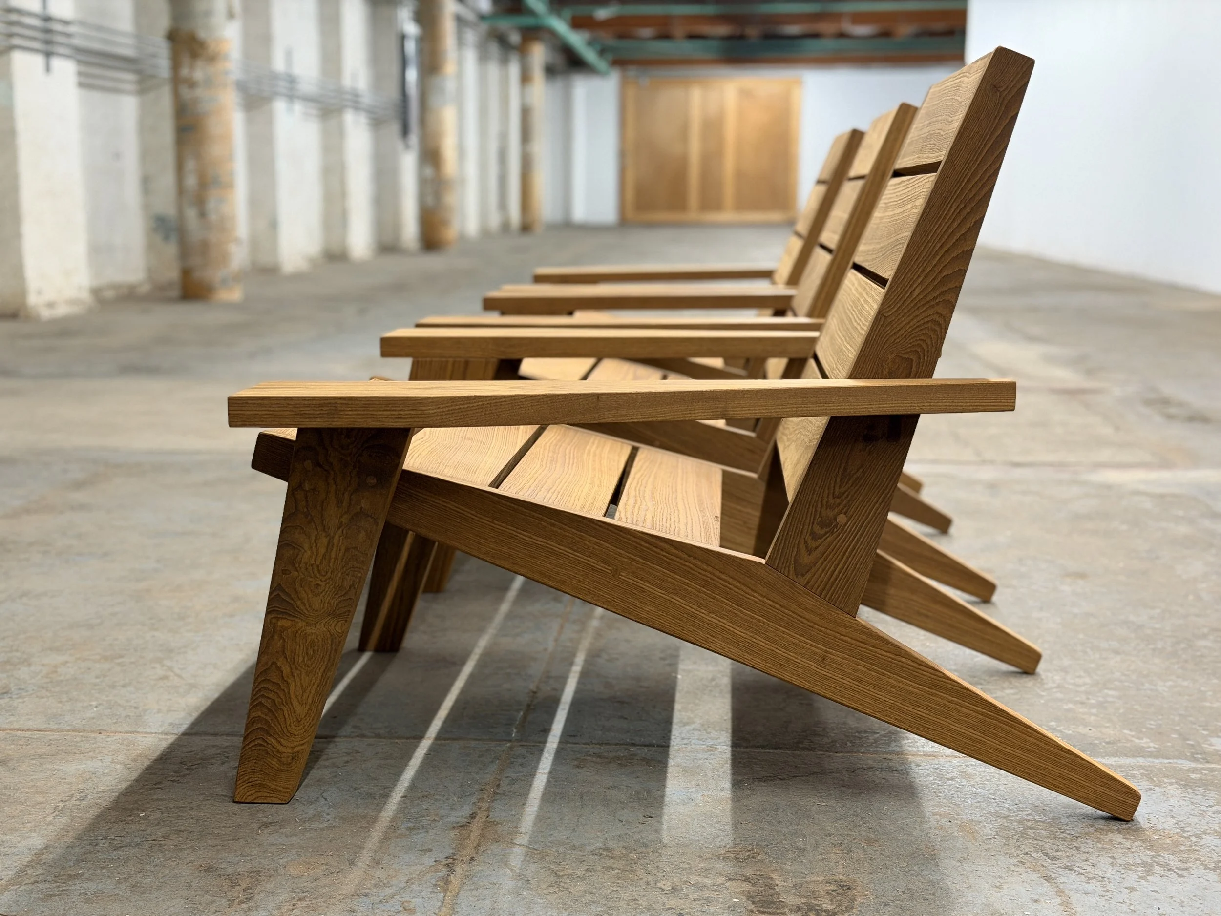 Multiple wooden outdoor lounge chairs lined up on a concrete floor in an indoor setting with wooden posts and a white wall.