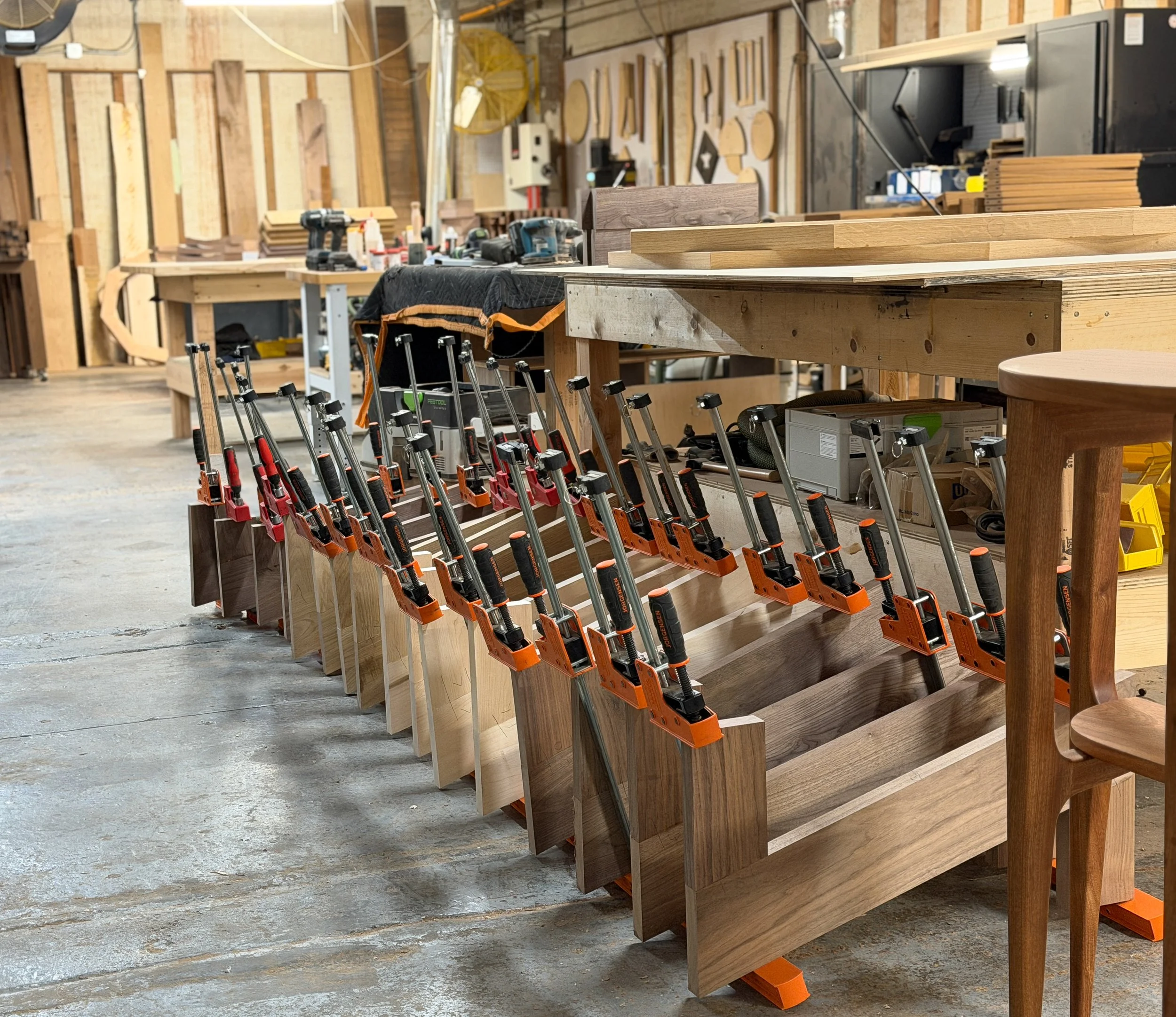 A woodworking workshop with clamps holding pieces of wood in a row, workbenches, tools, and woodwork in progress.