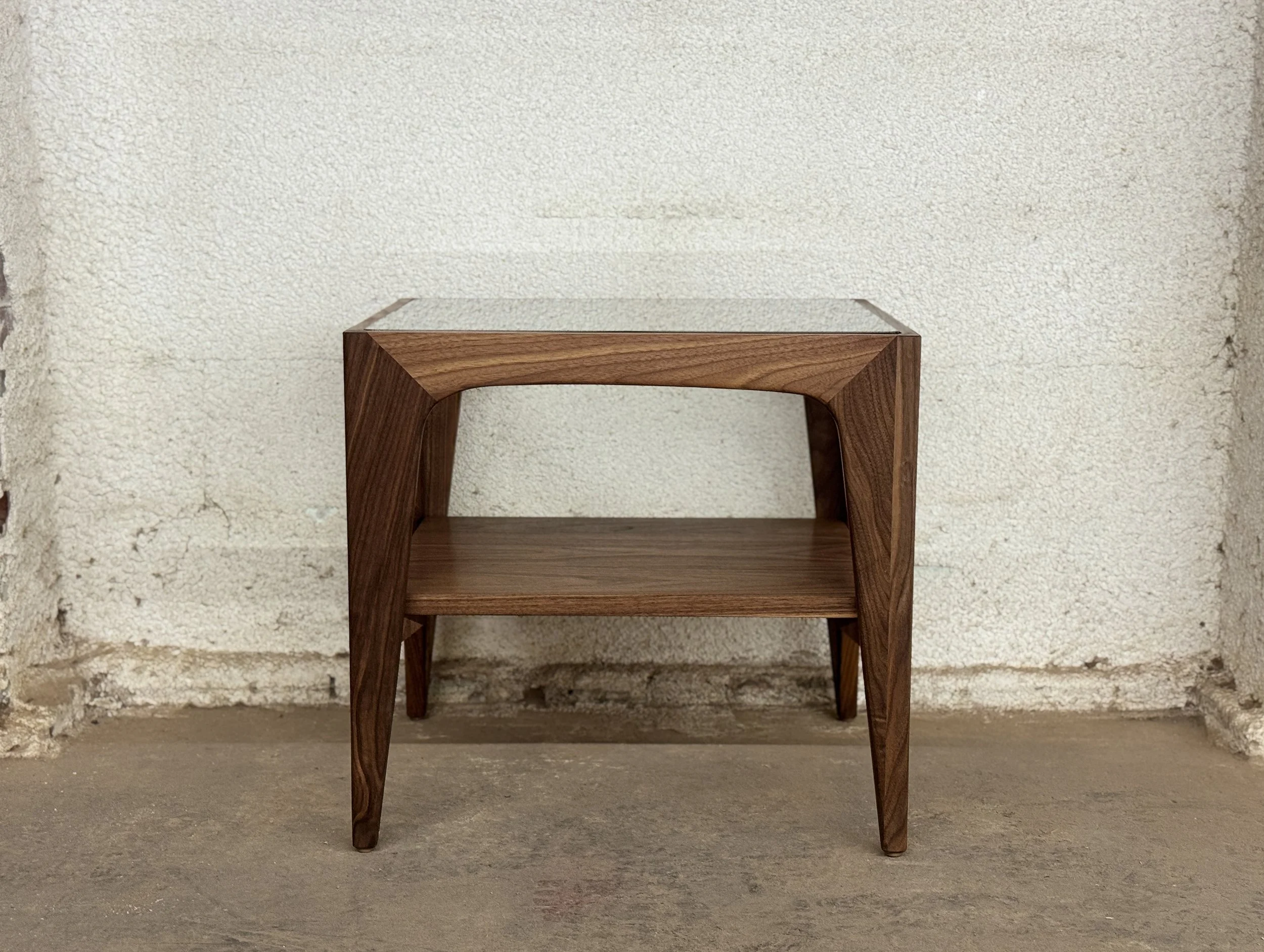 A modern wooden side table with a rectangular glass top, placed against a textured off-white wall and on a concrete floor.